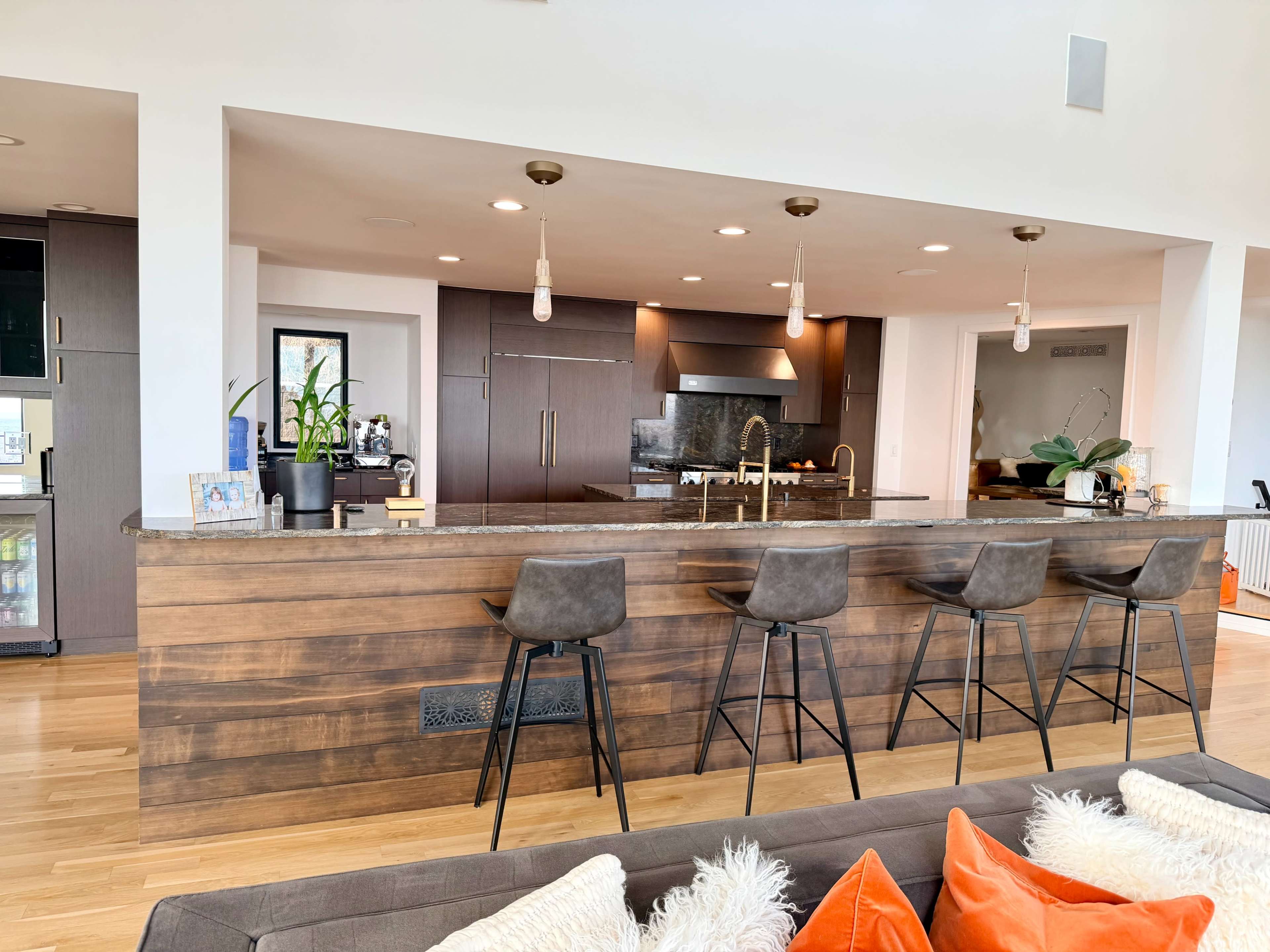 A modern kitchen features a large island with four bar stools, dark cabinets, and sleek appliances, illuminated by pendant lights.