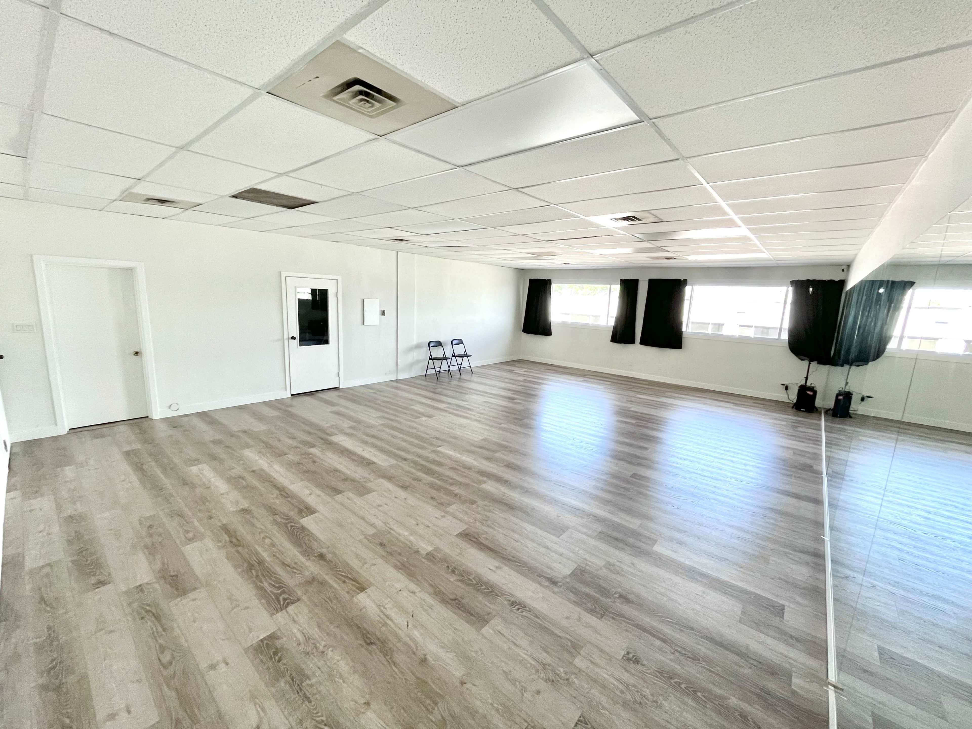 A spacious, empty room with wooden flooring, two doors, large windows with curtains, and a mirrored wall.