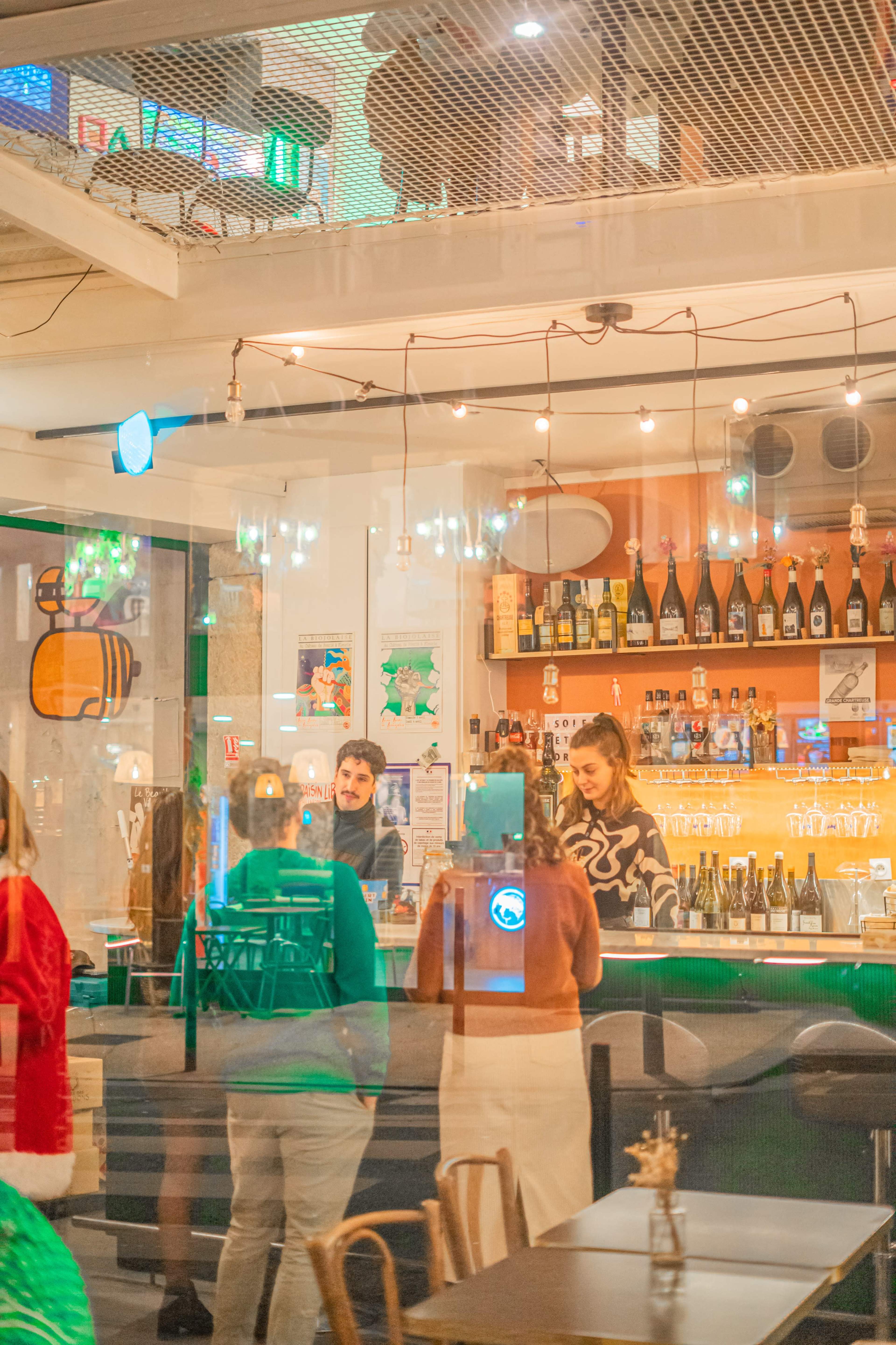 A bartender serves drinks at a lively bar while patrons interact in a warmly lit space filled with bottles and decorative lights.