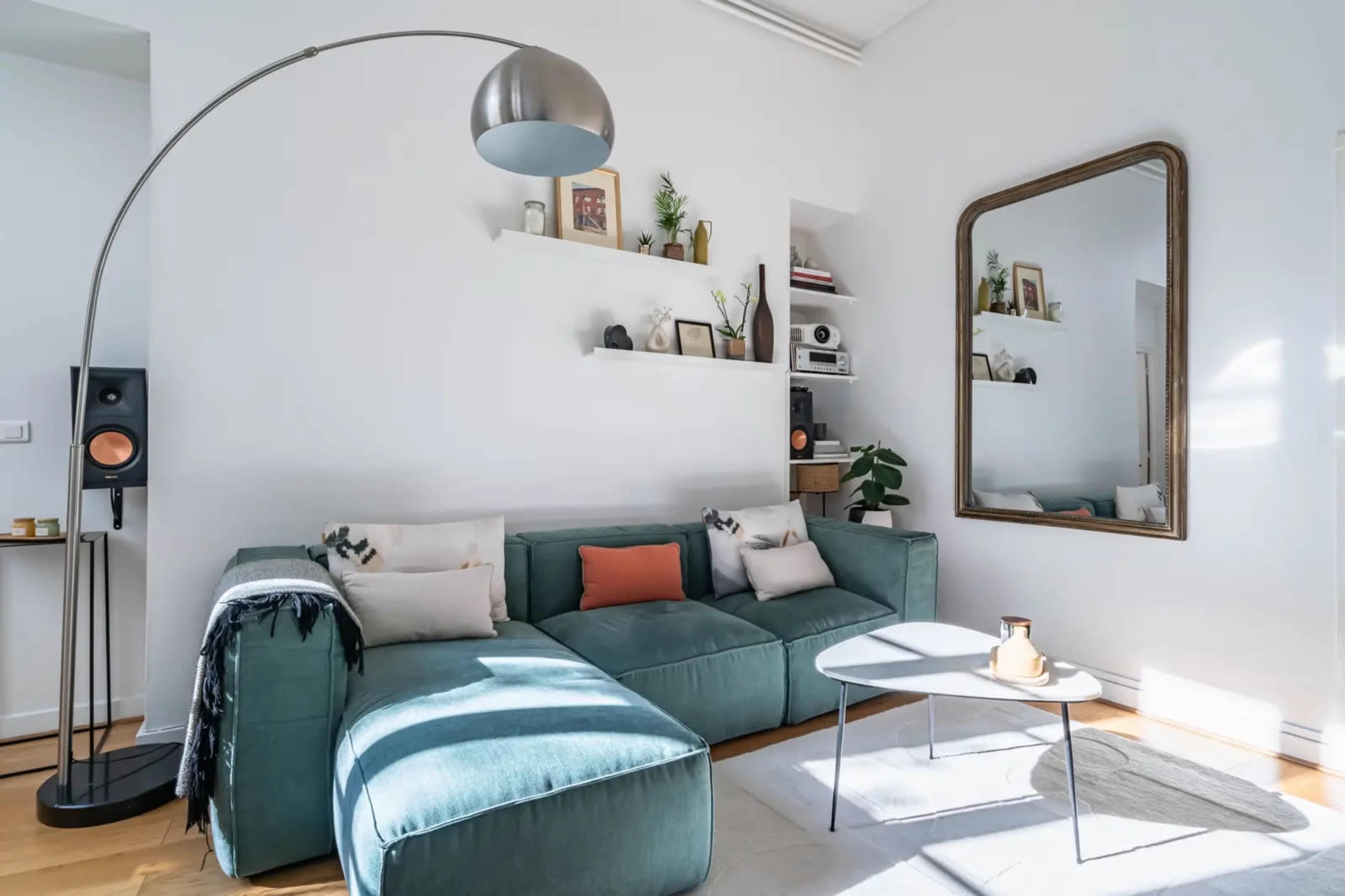 A modern living room features a sectional sofa, a large mirror, a floor lamp, and decorative shelves on a white wall.