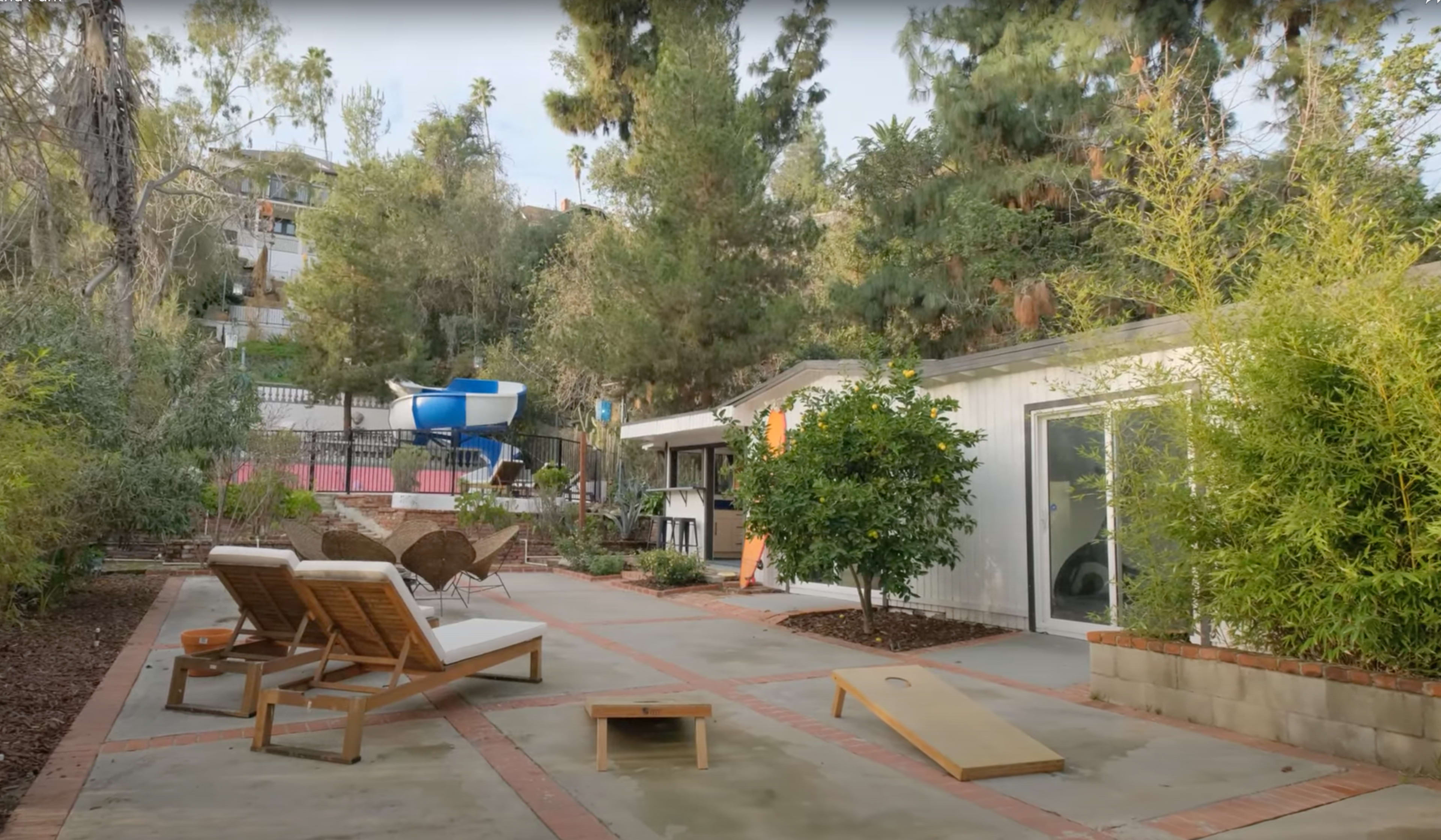 The scene features a backyard with concrete flooring, lounge chairs, a small ramp, and a water slide amid greenery and surrounding trees.
