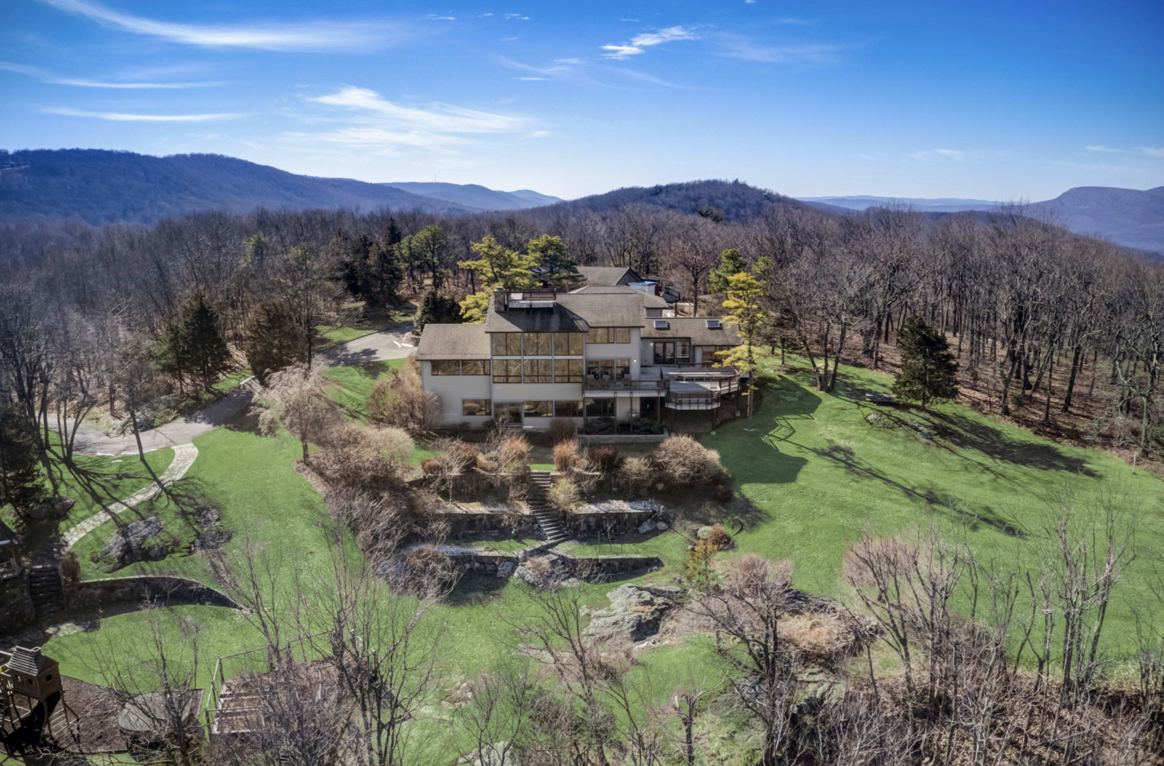A large, modern house sits on a green hillside surrounded by sparse trees and distant mountains under a clear blue sky.