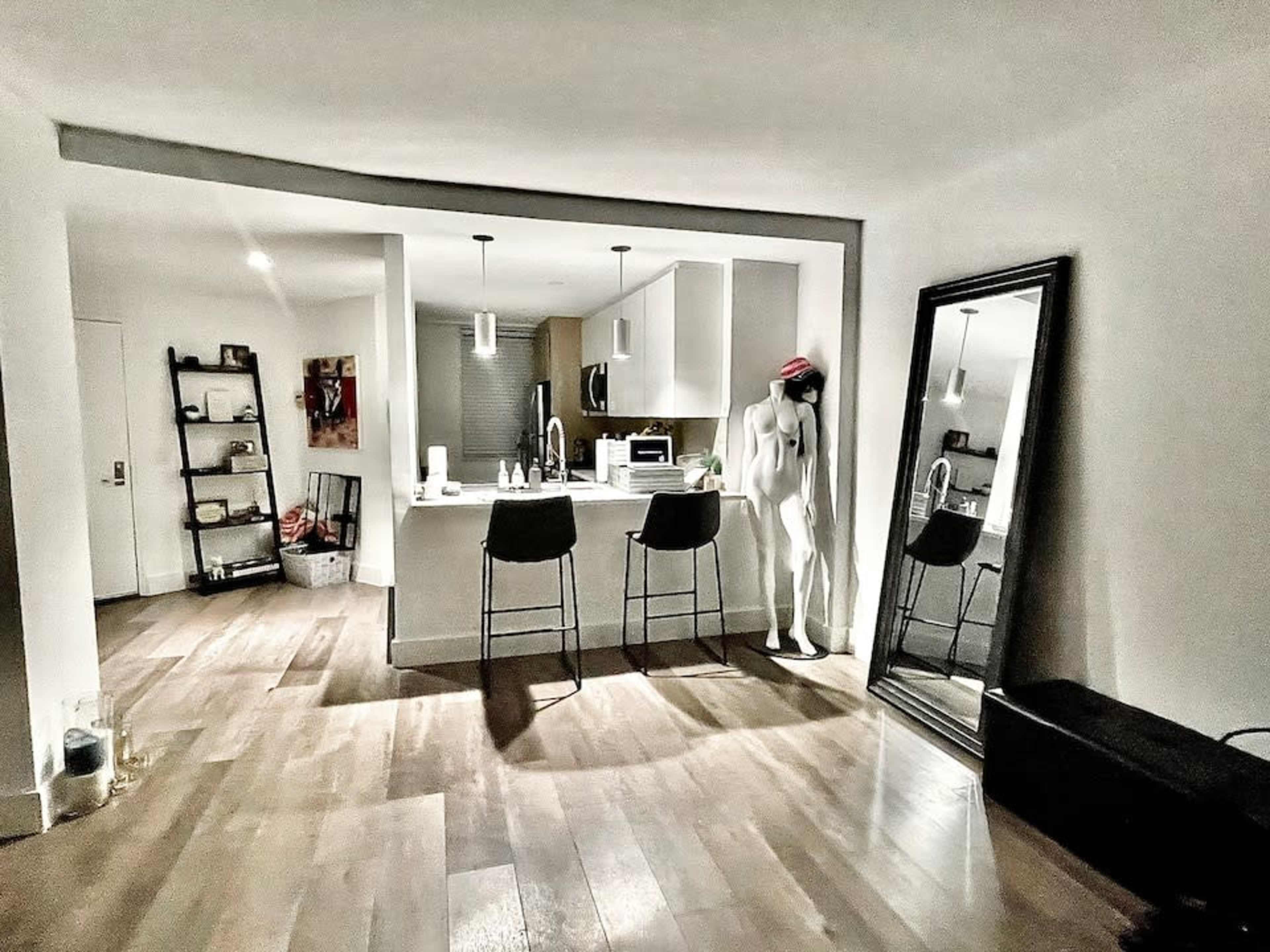 A modern kitchen with a central island, bar stools, a mannequin wearing a red hat, and a large mirror reflecting the surroundings.
