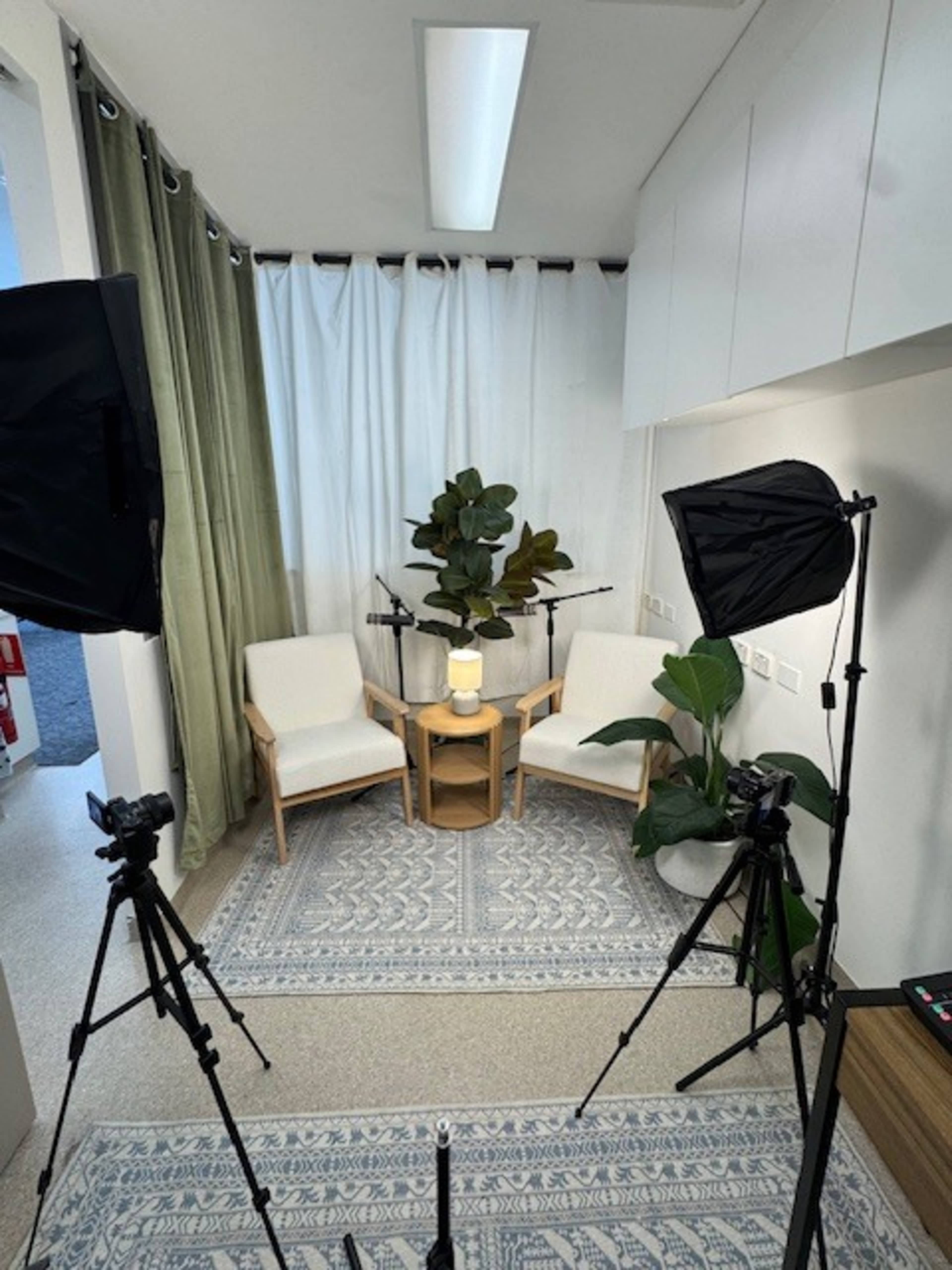 The image shows a small studio setup with two chairs, a low table, two camera tripods with cameras, and decorative plants against a backdrop of curtains.