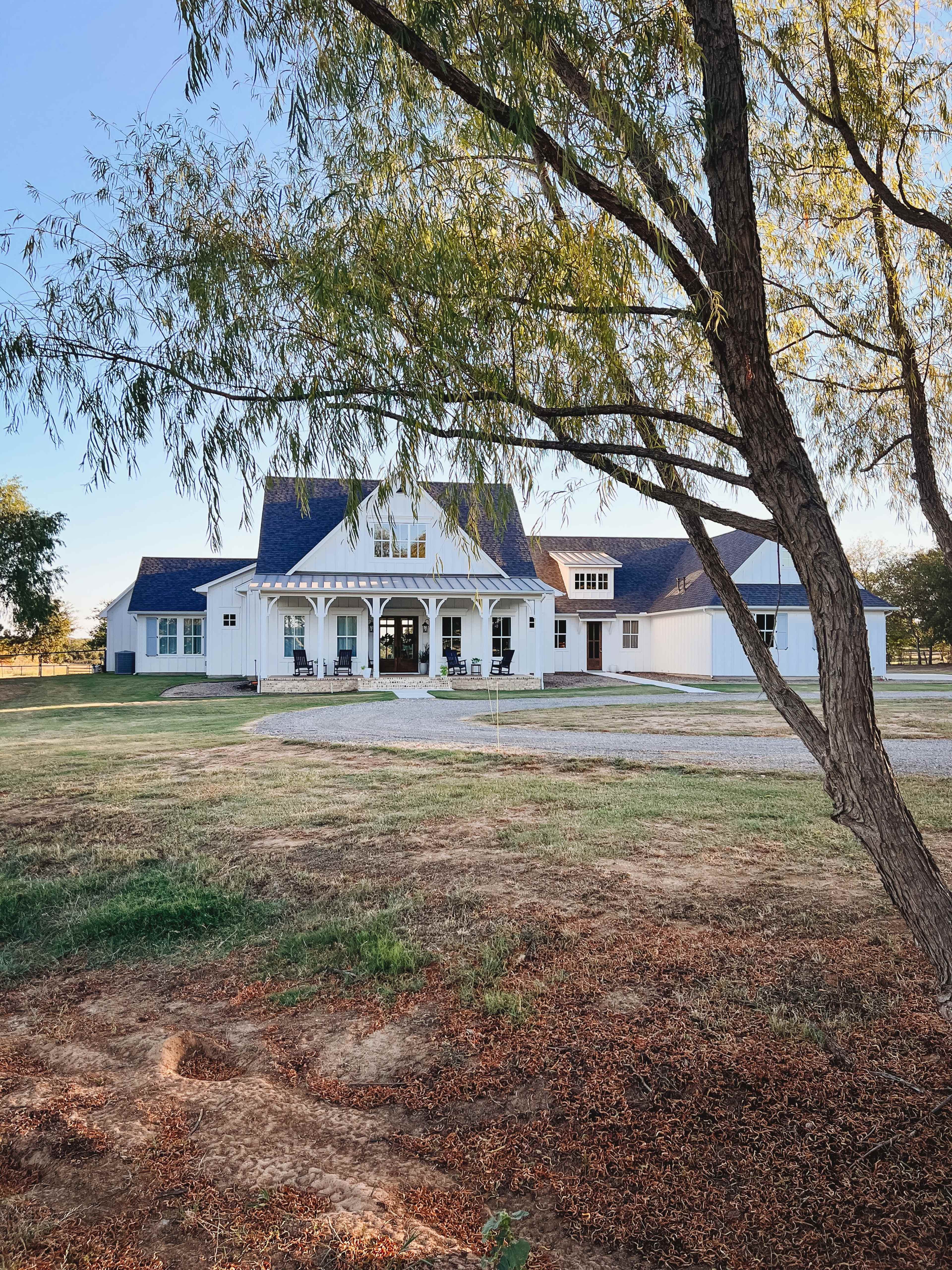 A white farmhouse with a large porch and a sloped roof is set against a grassy landscape, bordered by trees.
