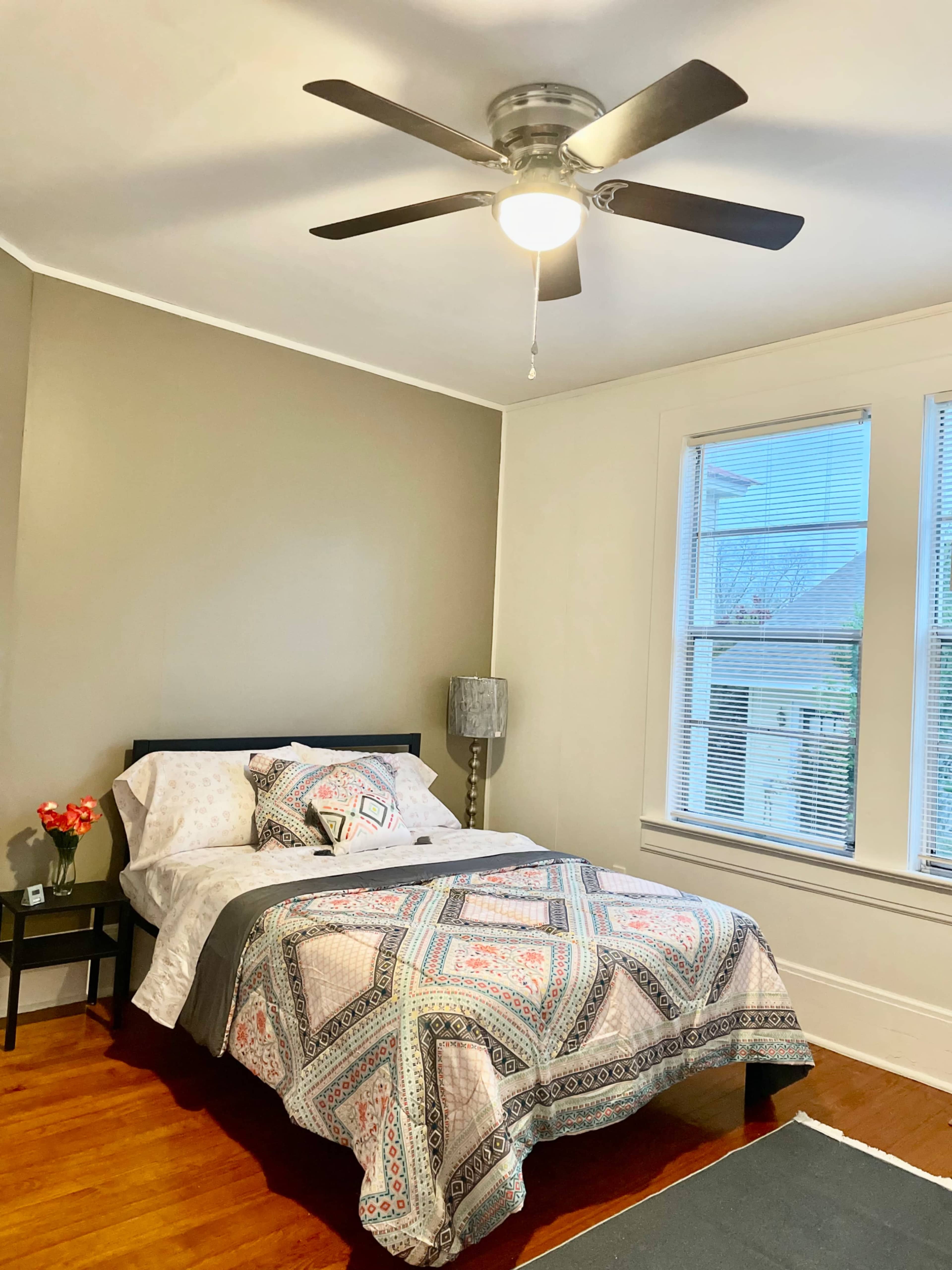 The image shows a bedroom with a bed dressed in patterned bedding, a nightstand with a lamp, and a ceiling fan above.