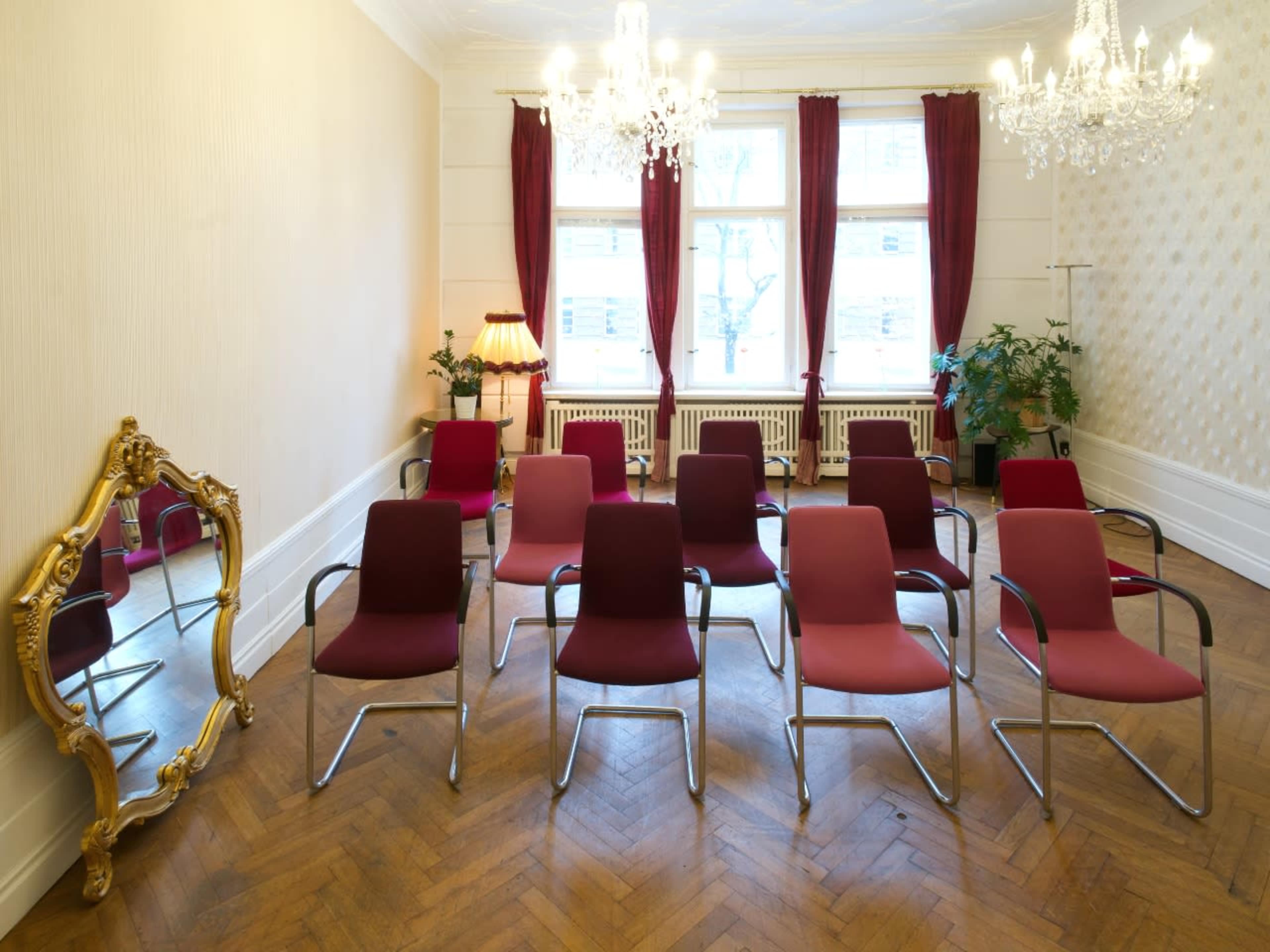 A room with a row of red chairs arranged facing a large mirror, with chandeliers hanging from the ceiling and a lamp in the corner.