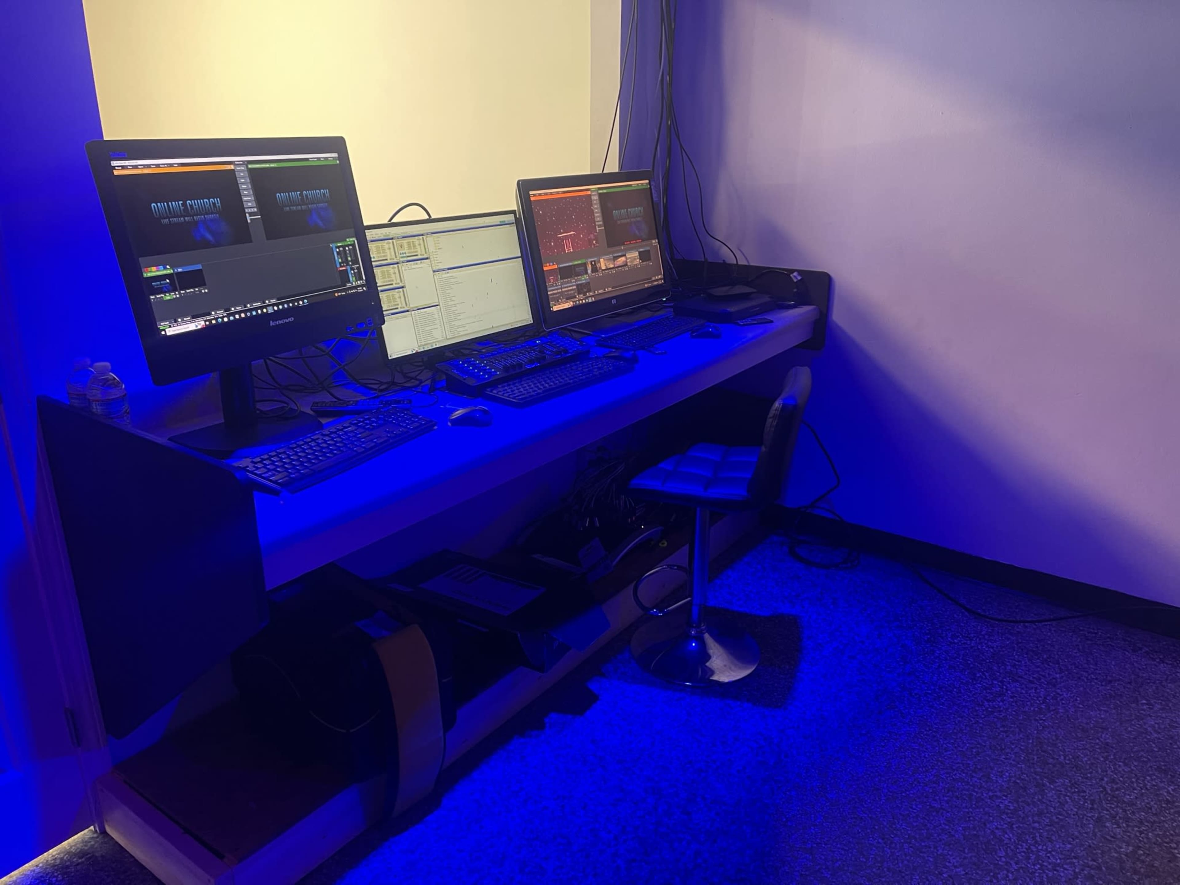 A modern workstation with three computer monitors, keyboards, and a chair, set against a wall illuminated by blue light.