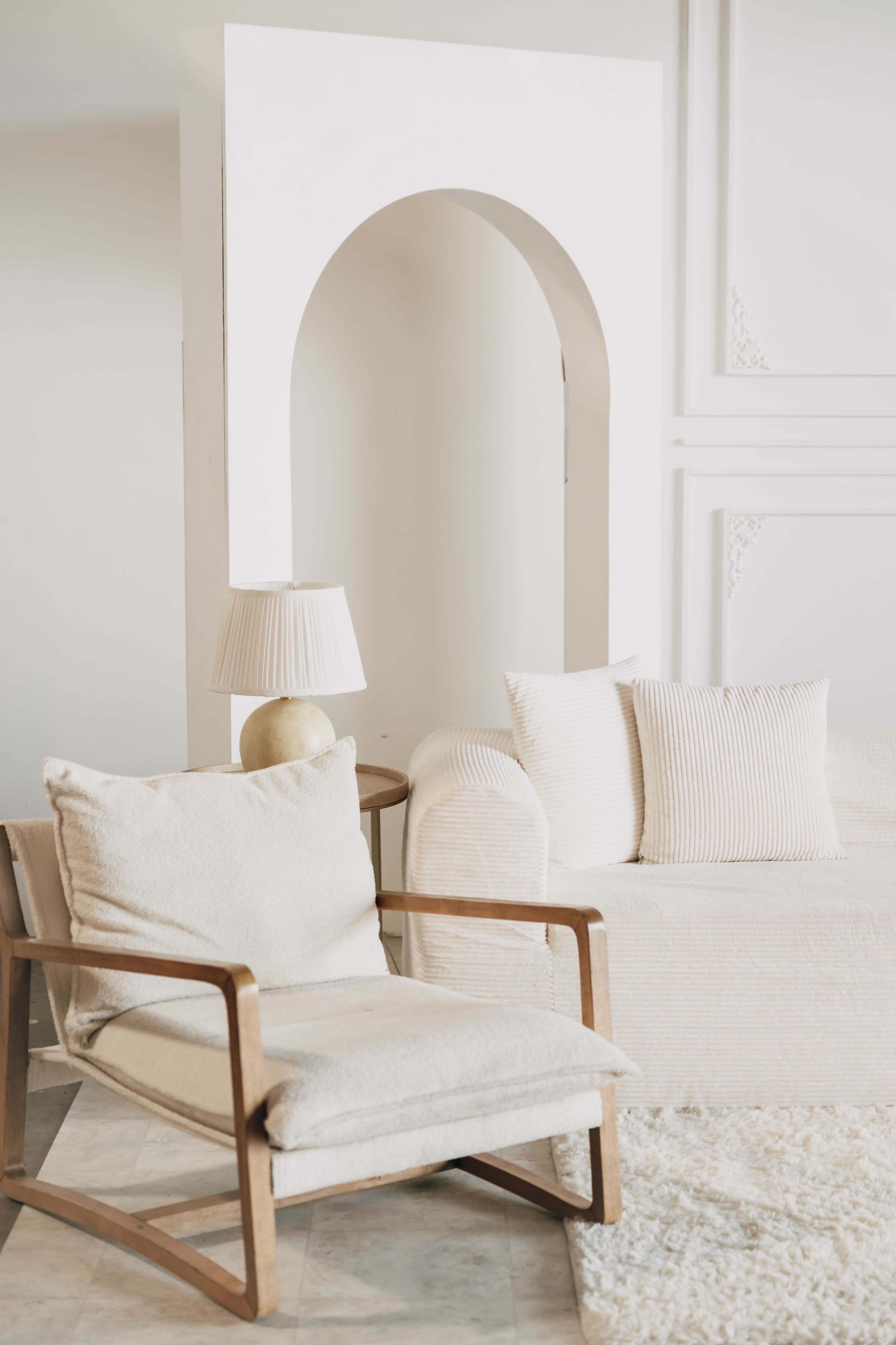 The image shows a light and airy living space featuring a white sofa, a cream-colored armchair, and a decorative archway in the background.