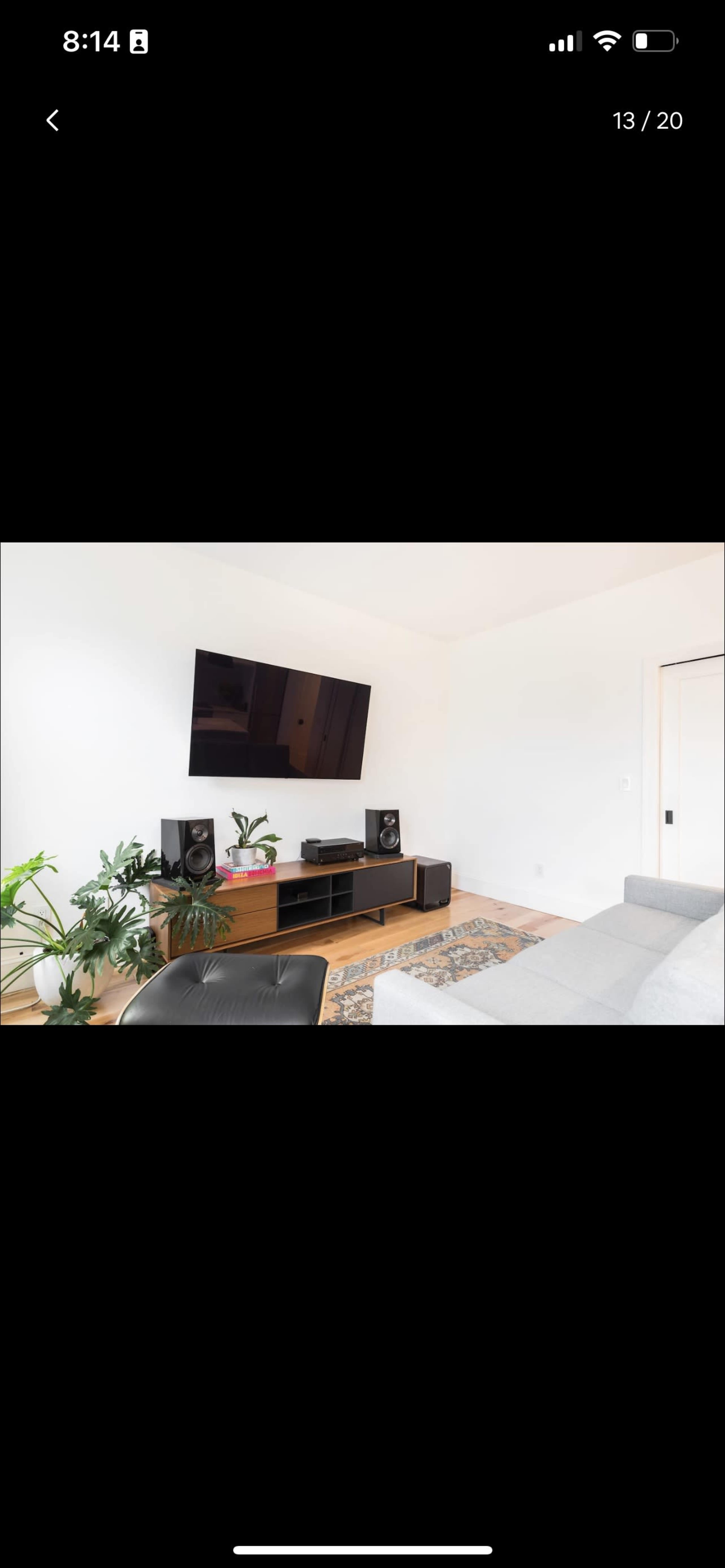 The image shows a modern living room with a wall-mounted television above a wooden entertainment unit and a gray sofa, accompanied by houseplants.