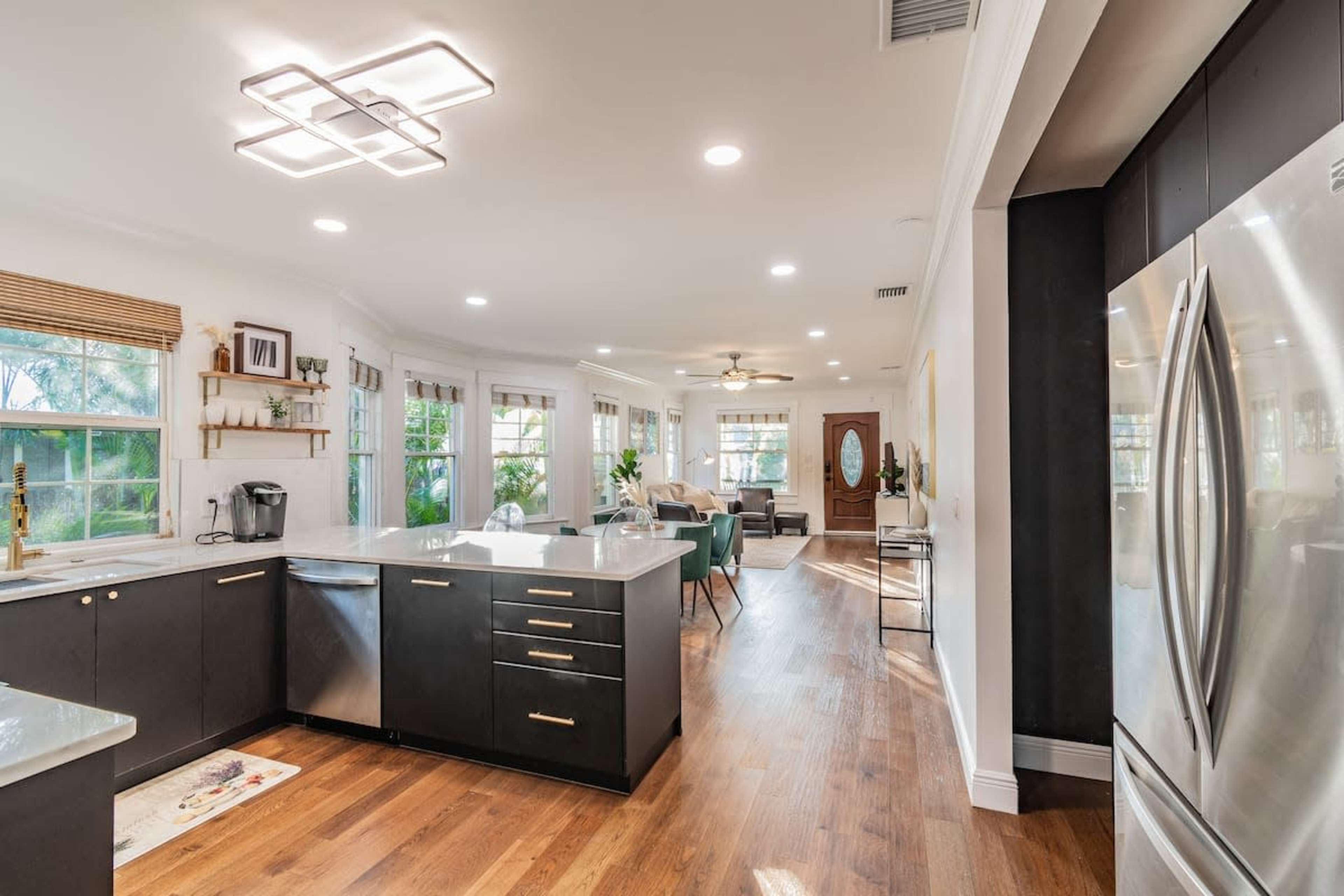 A modern kitchen with dark cabinetry and a breakfast bar that opens into a bright living area featuring large windows and comfortable seating.