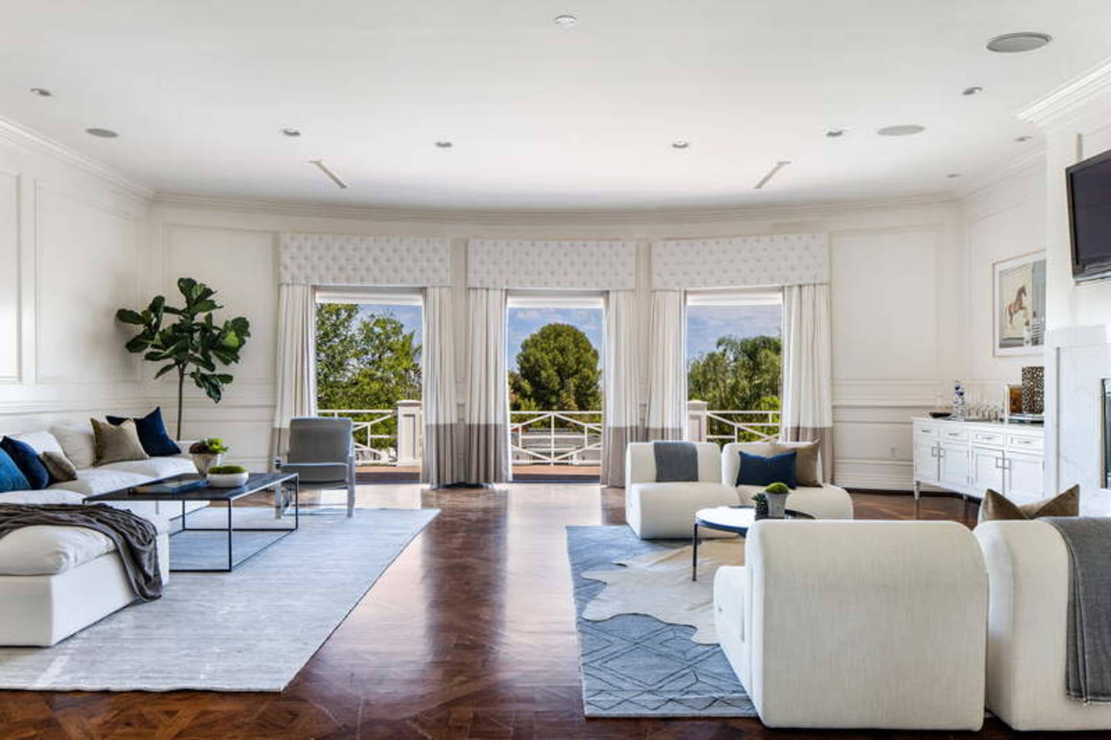 A spacious living room features large windows with views of greenery, a sectional sofa, armchairs, and a television mounted on the wall.