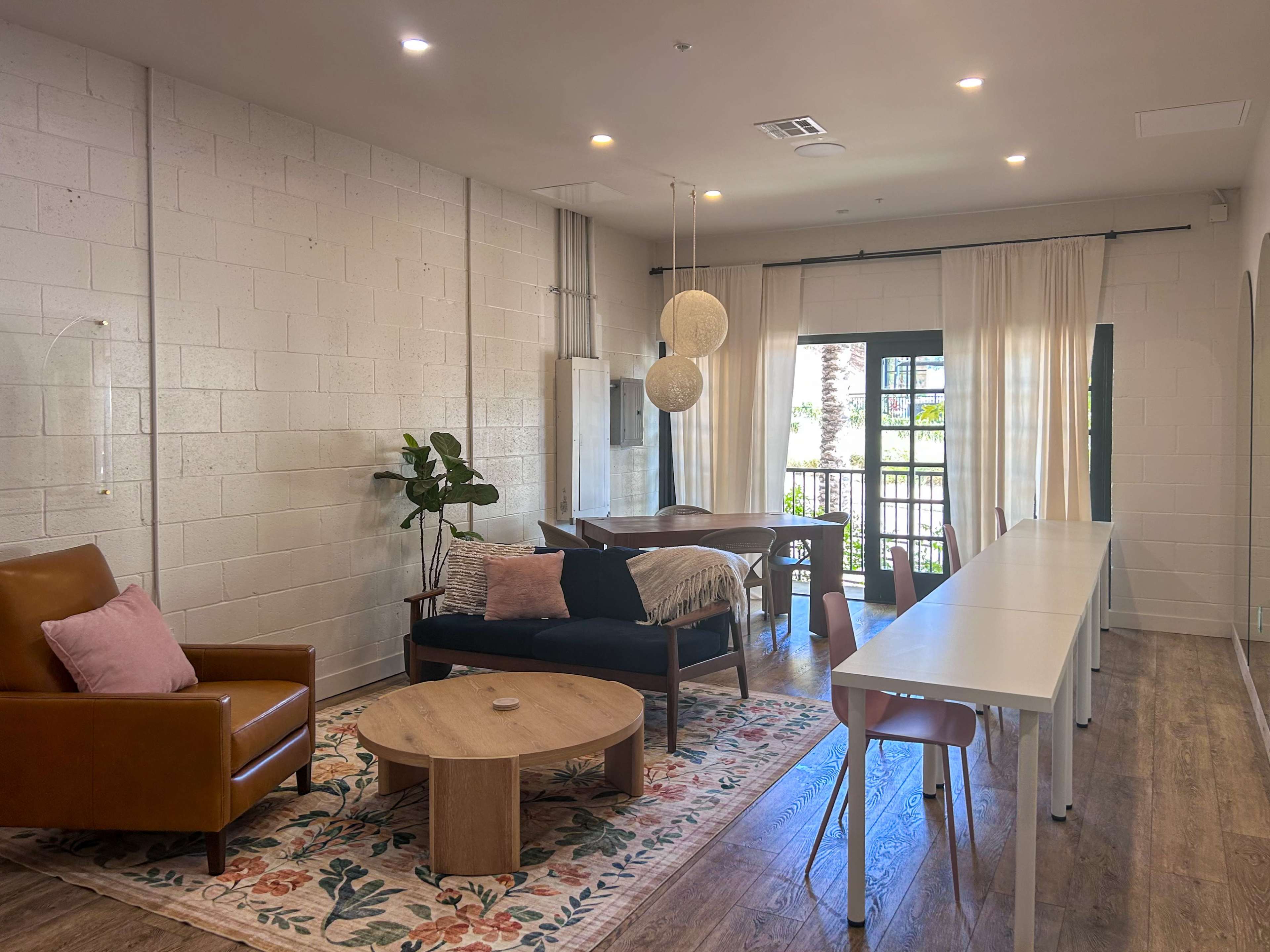 The image depicts a modern, well-lit lounge area featuring a leather armchair, a wooden coffee table, a long white communal table, and large windows with light curtains.