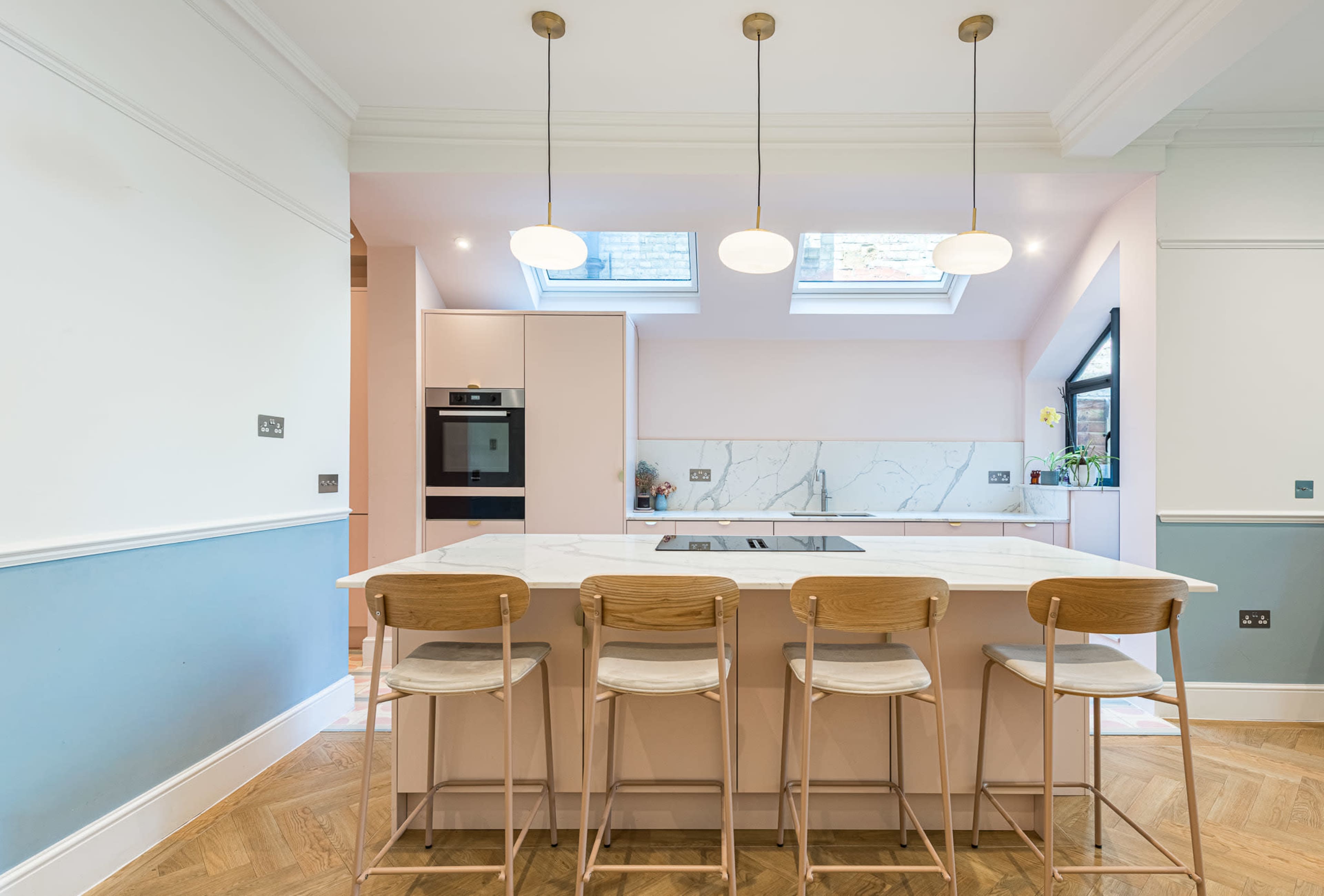 The image shows a modern kitchen with a central island, four wooden stools, and bright lighting fixtures above.