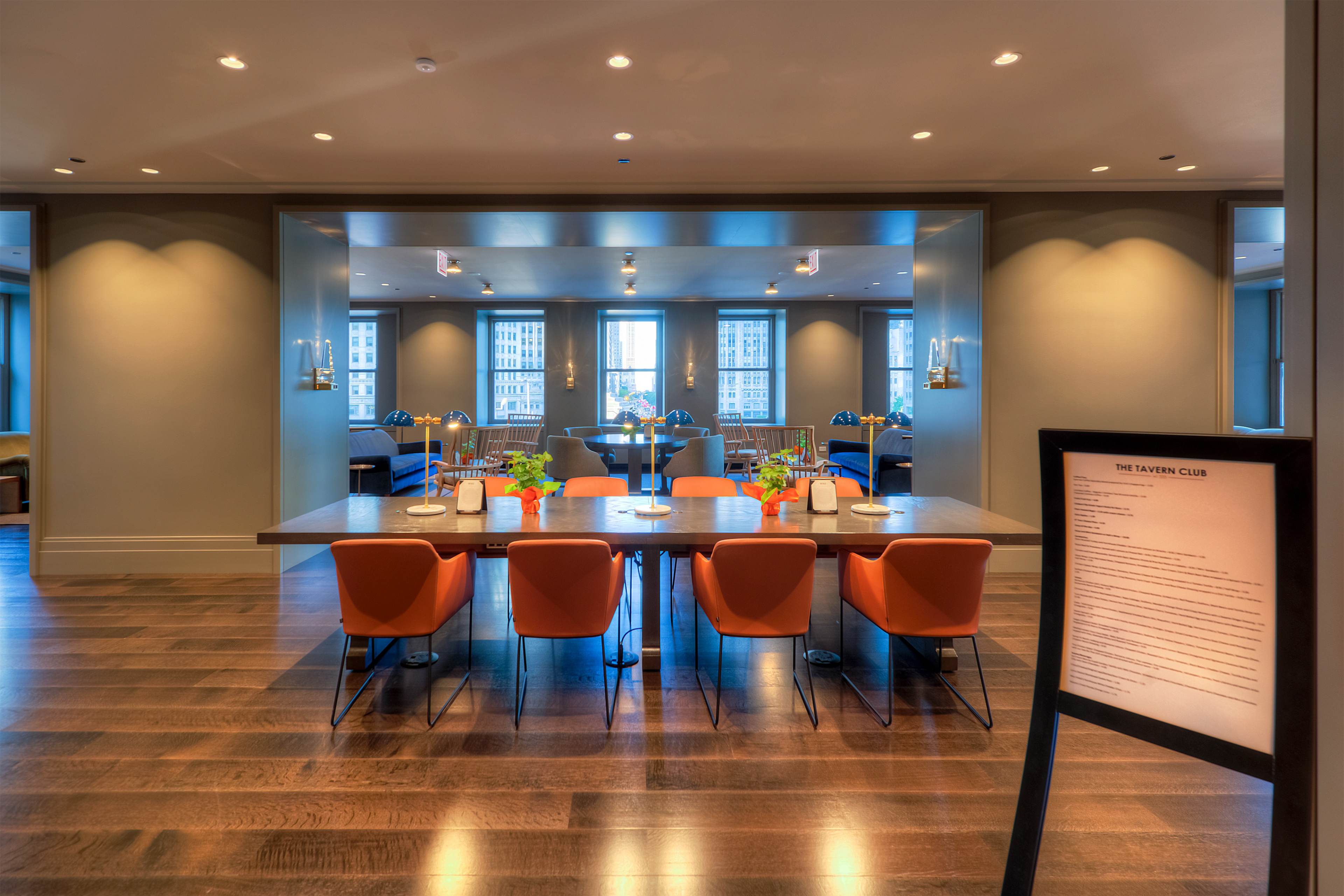 A modern dining area features a long table surrounded by orange chairs, with large windows providing a view of the outside.