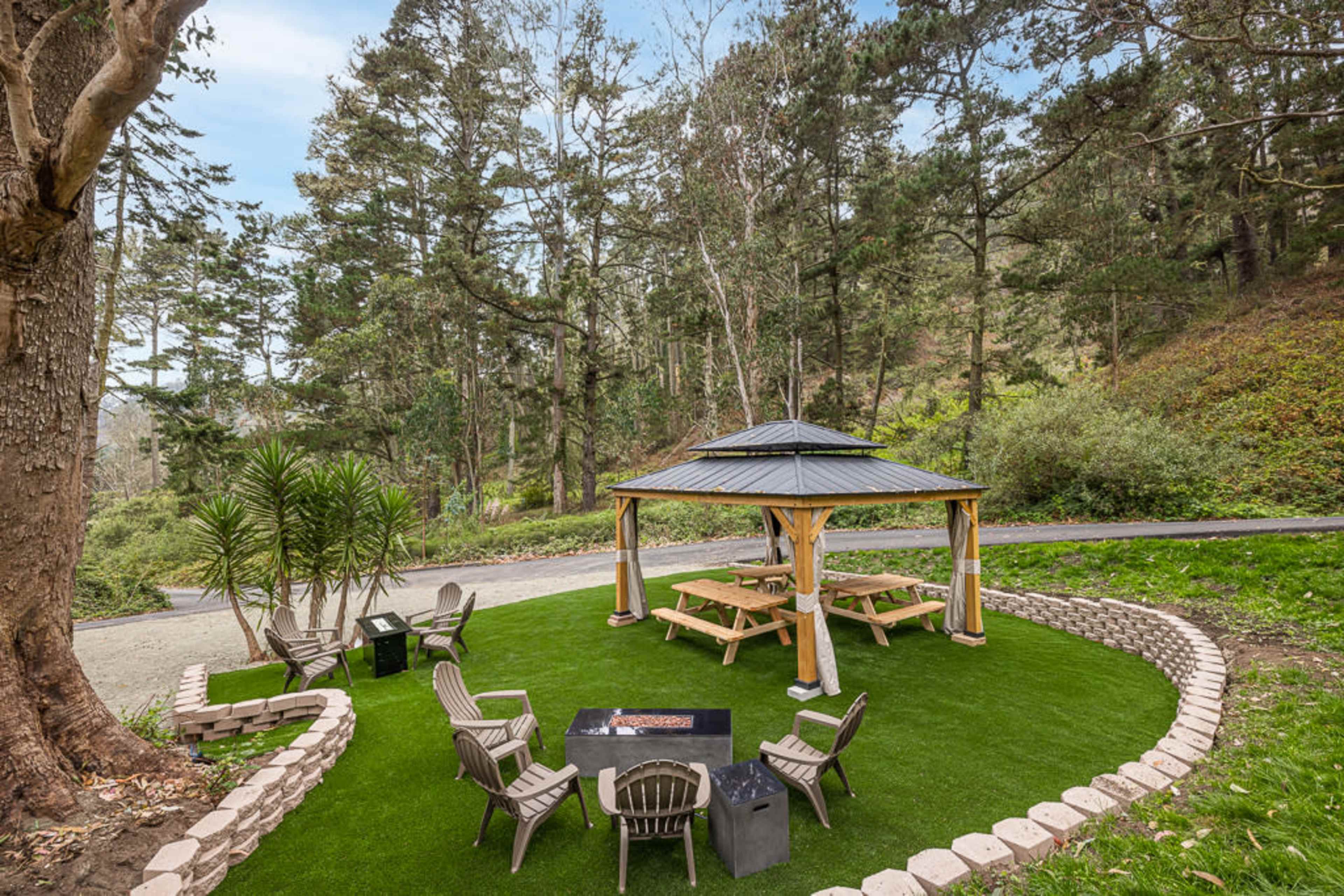 A gazebo with picnic tables and seating surrounded by landscaped grass and trees in a wooded area.