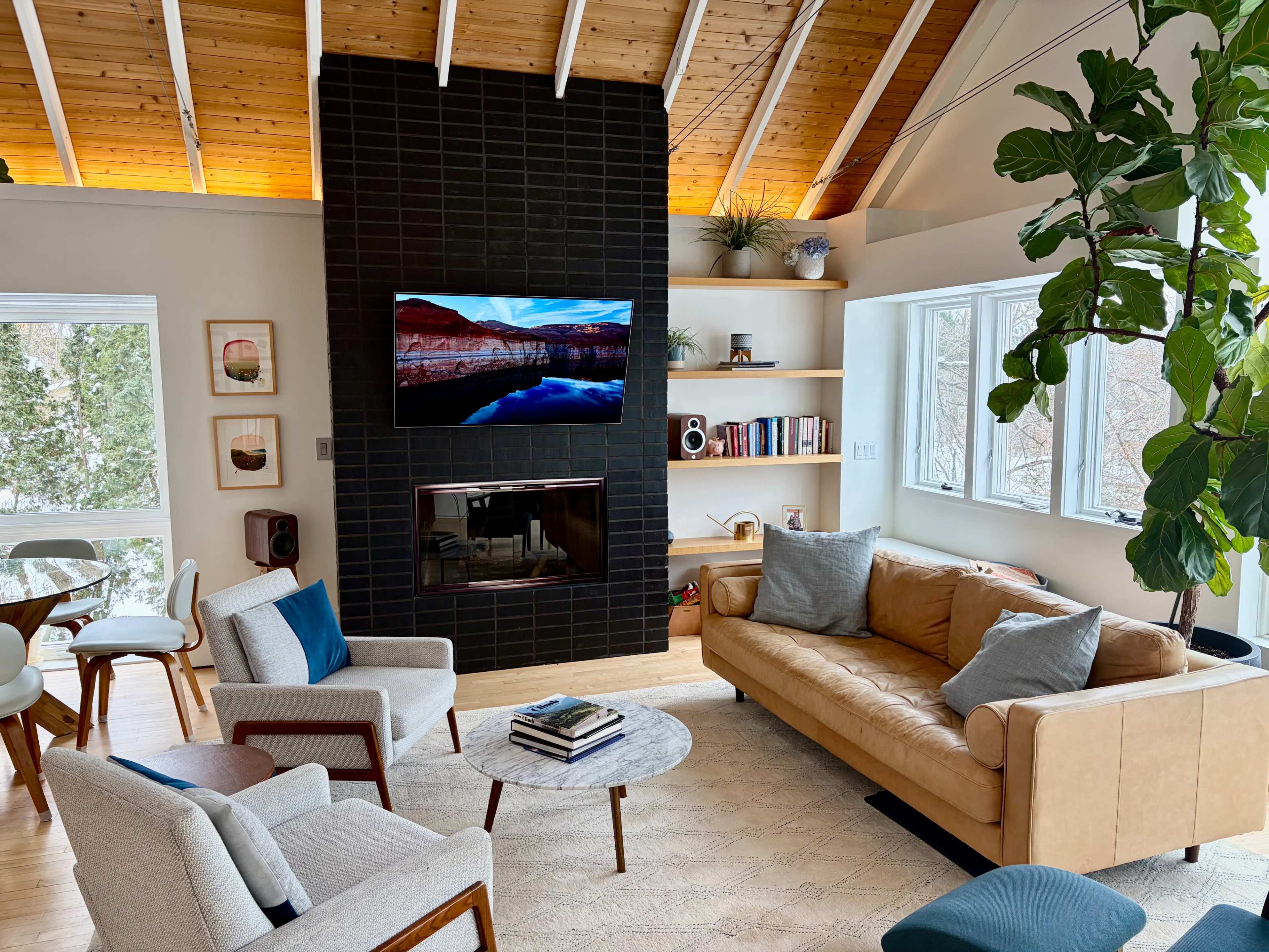 The image shows a modern living room featuring a brown leather couch, two armchairs, a round coffee table, and a mounted television above a fireplace, with large windows and wooden ceiling beams.