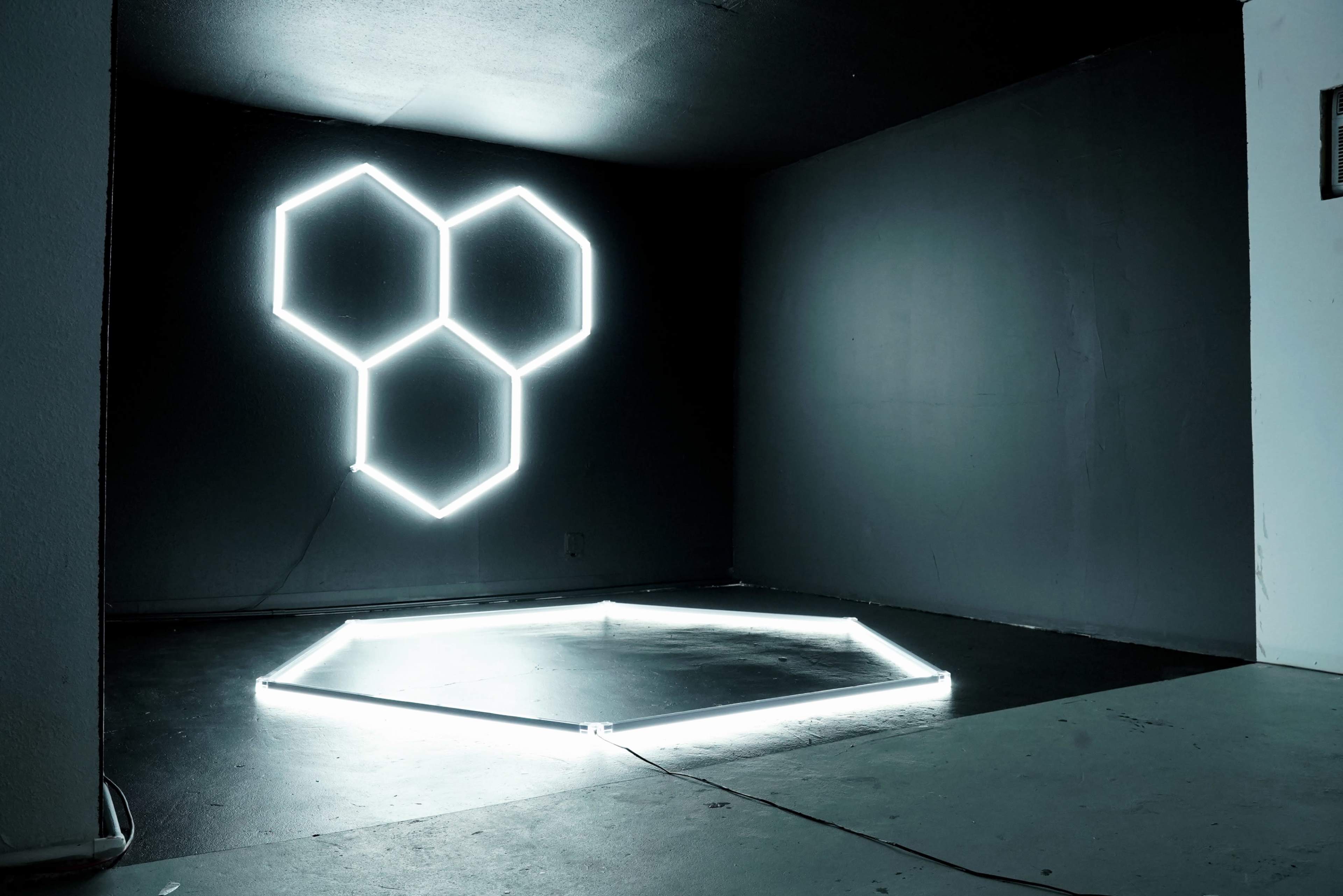 The image shows a dimly lit room with a wall featuring a hexagonal neon light arrangement and a matching hexagonal outline on the floor.