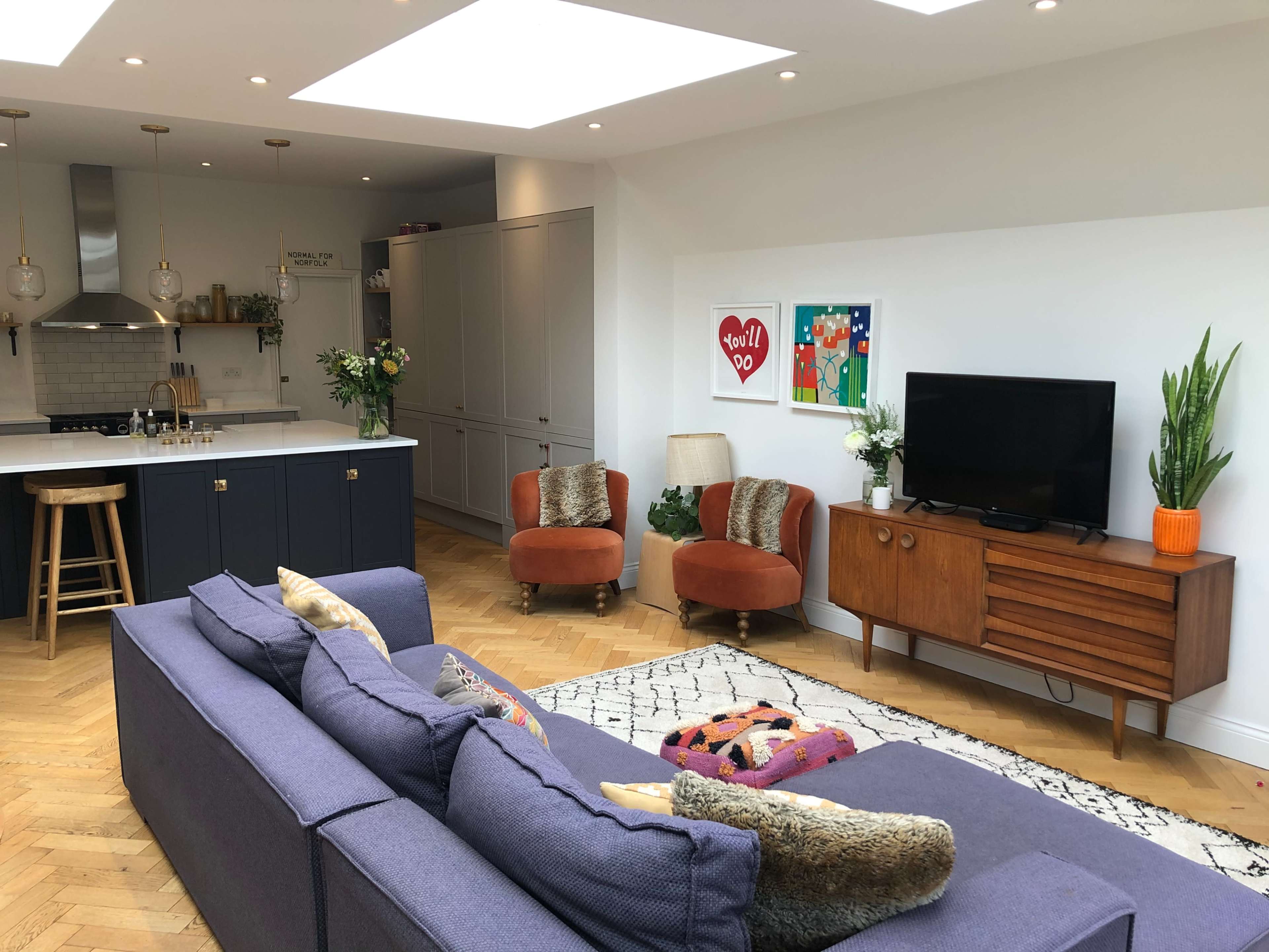 A modern living room features a blue sofa facing a wooden television stand, with two orange chairs, a coffee table, and a kitchen area in the background.
