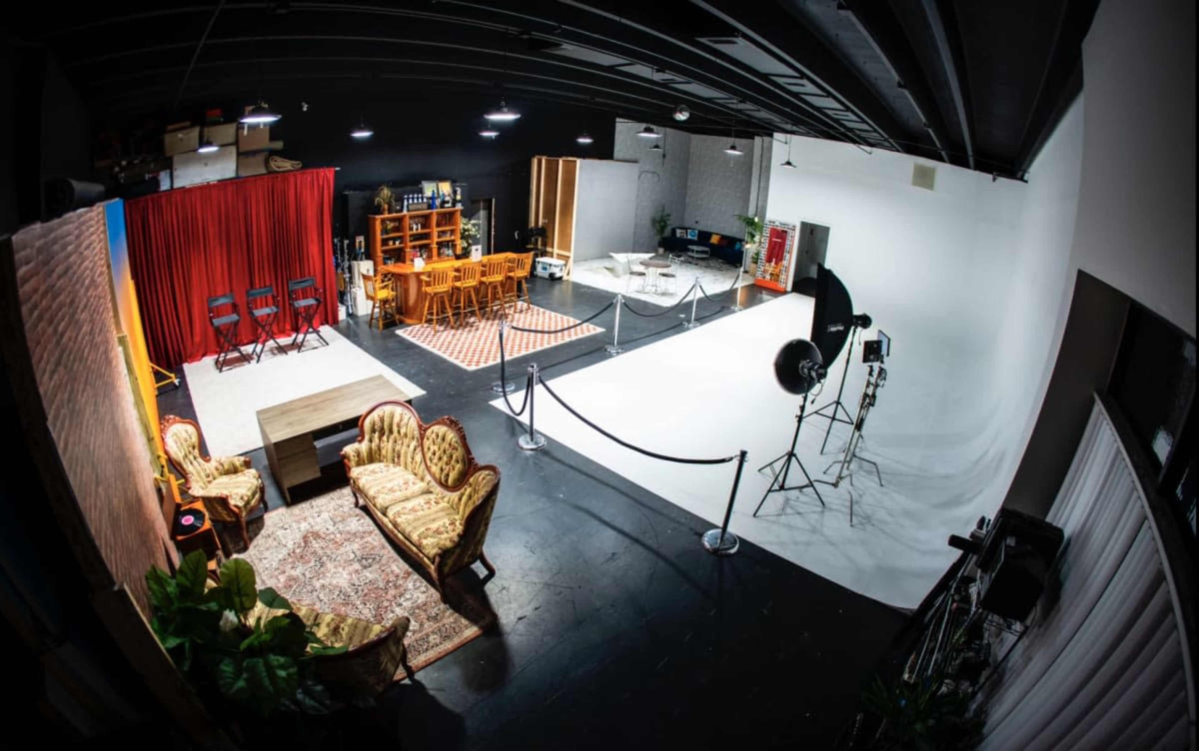 The image shows a spacious studio featuring a seating area with patterned sofas, a dining table and chairs, a white backdrop, and lighting equipment.