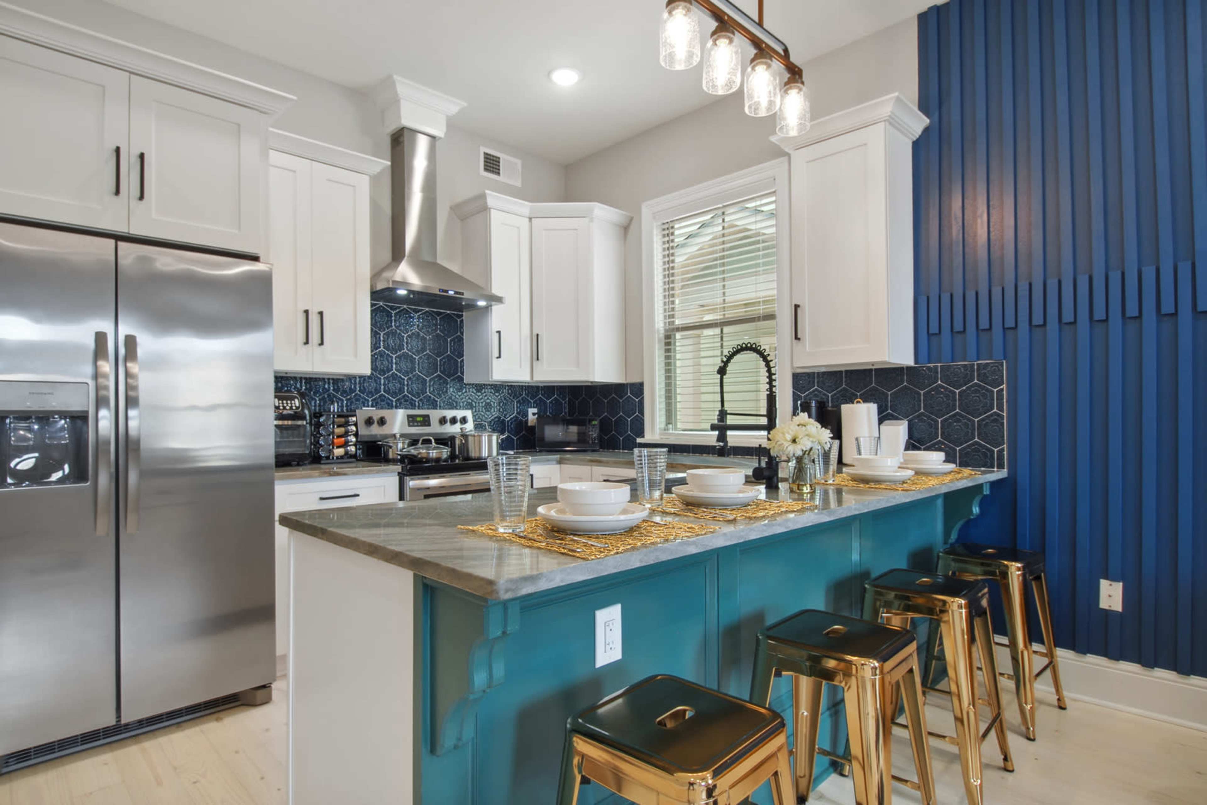 A modern kitchen features a central island with metallic bar stools, sleek cabinets, and a blue accent wall, complemented by geometric tile backsplash.