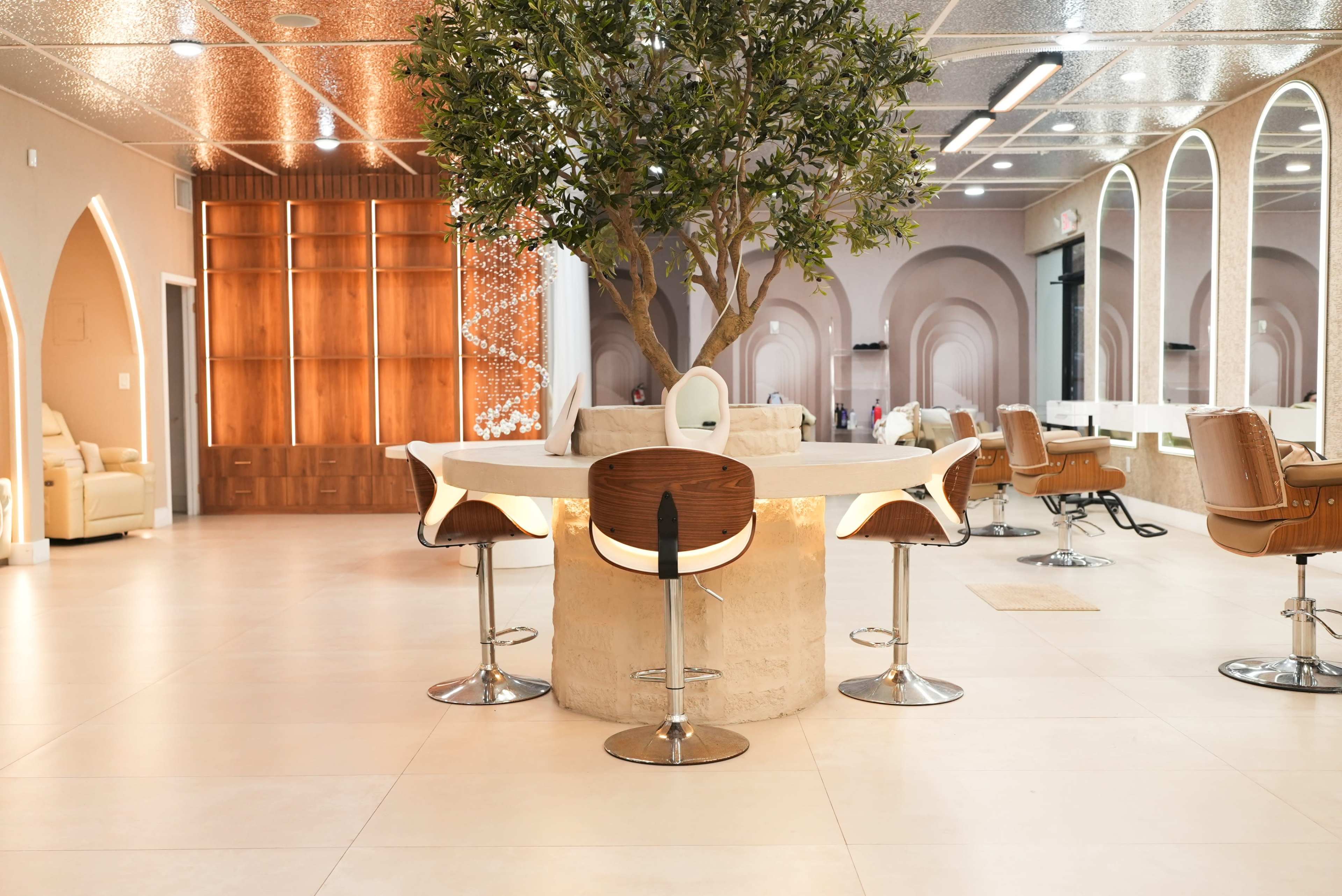 The image shows a modern salon interior featuring a central table with seating around it, accented by a decorative tree and large mirrors on the walls.