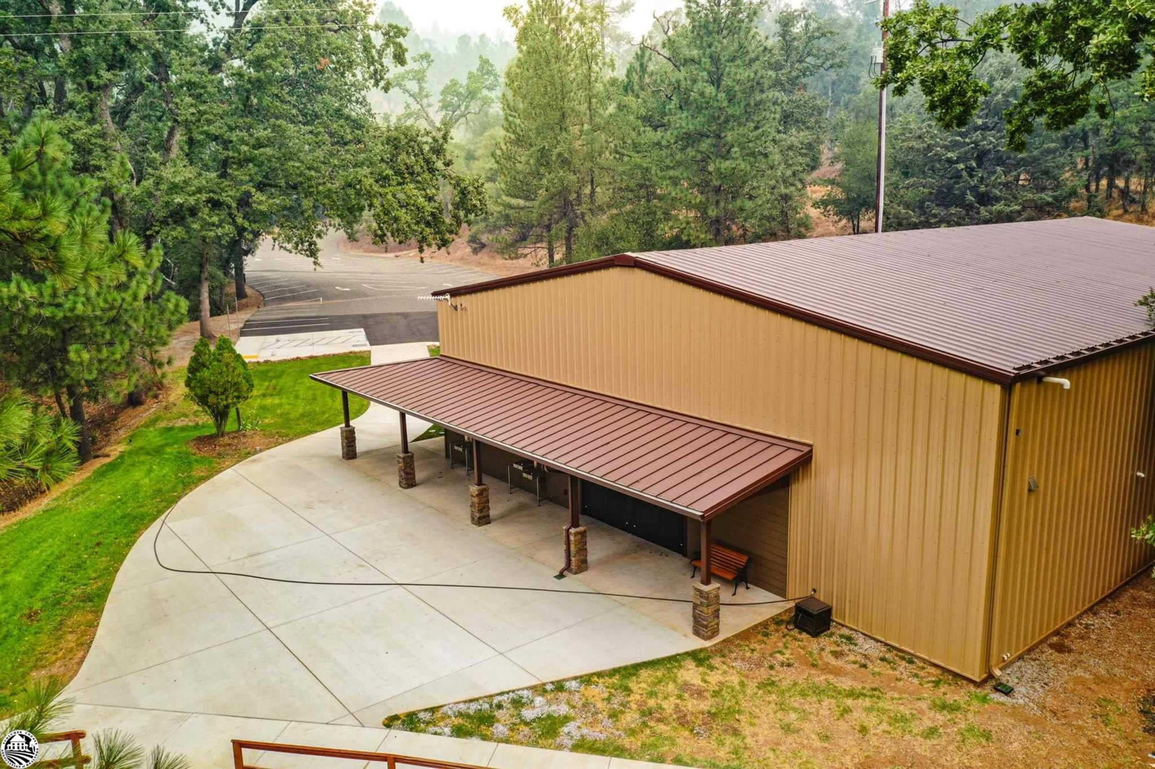 A large, tan metal building with a sloped roof, surrounded by trees and a concrete parking area.