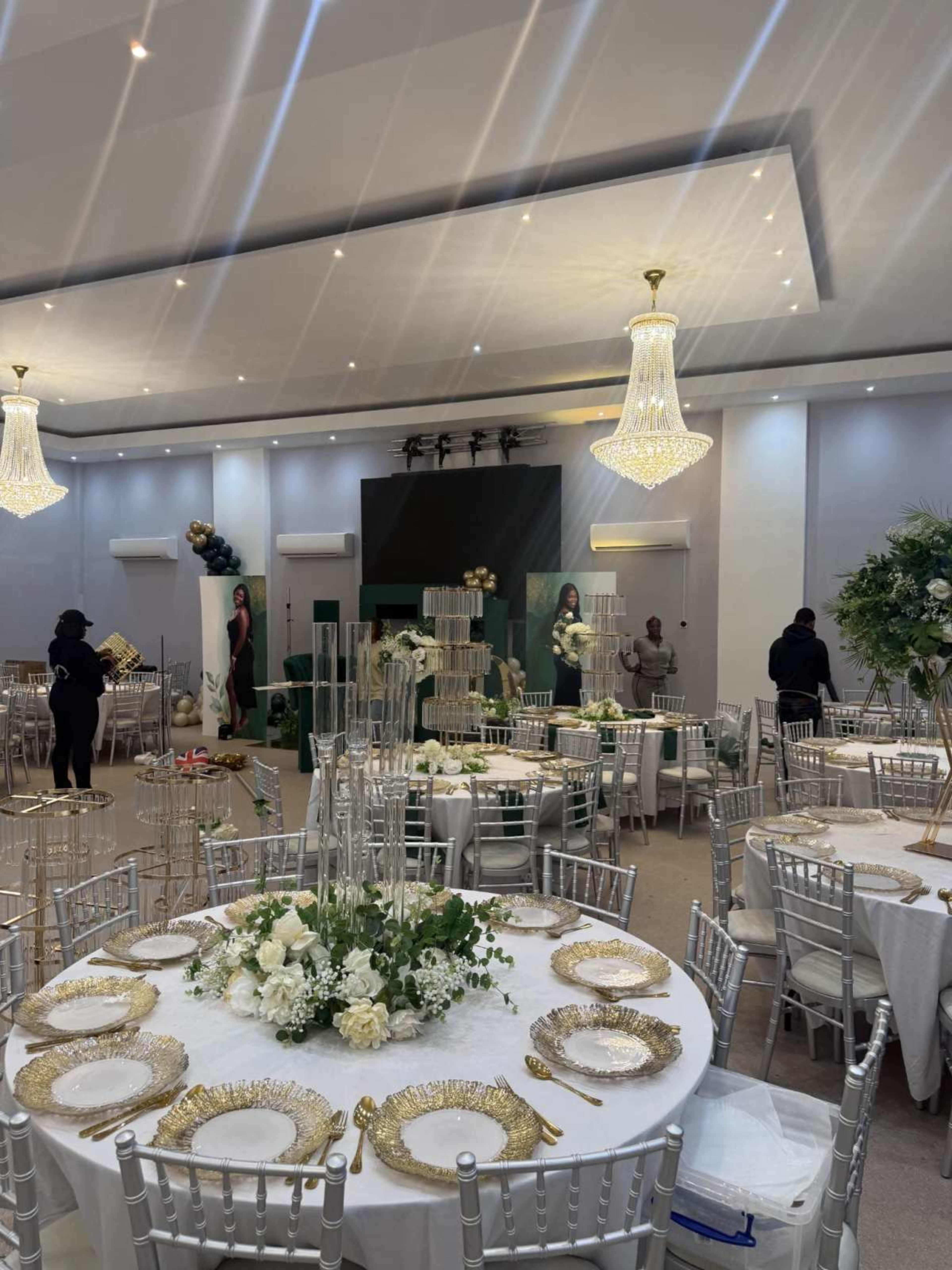 The image shows a banquet hall set up for an event, featuring elegantly dressed tables with floral centerpieces and large chandeliers overhead.