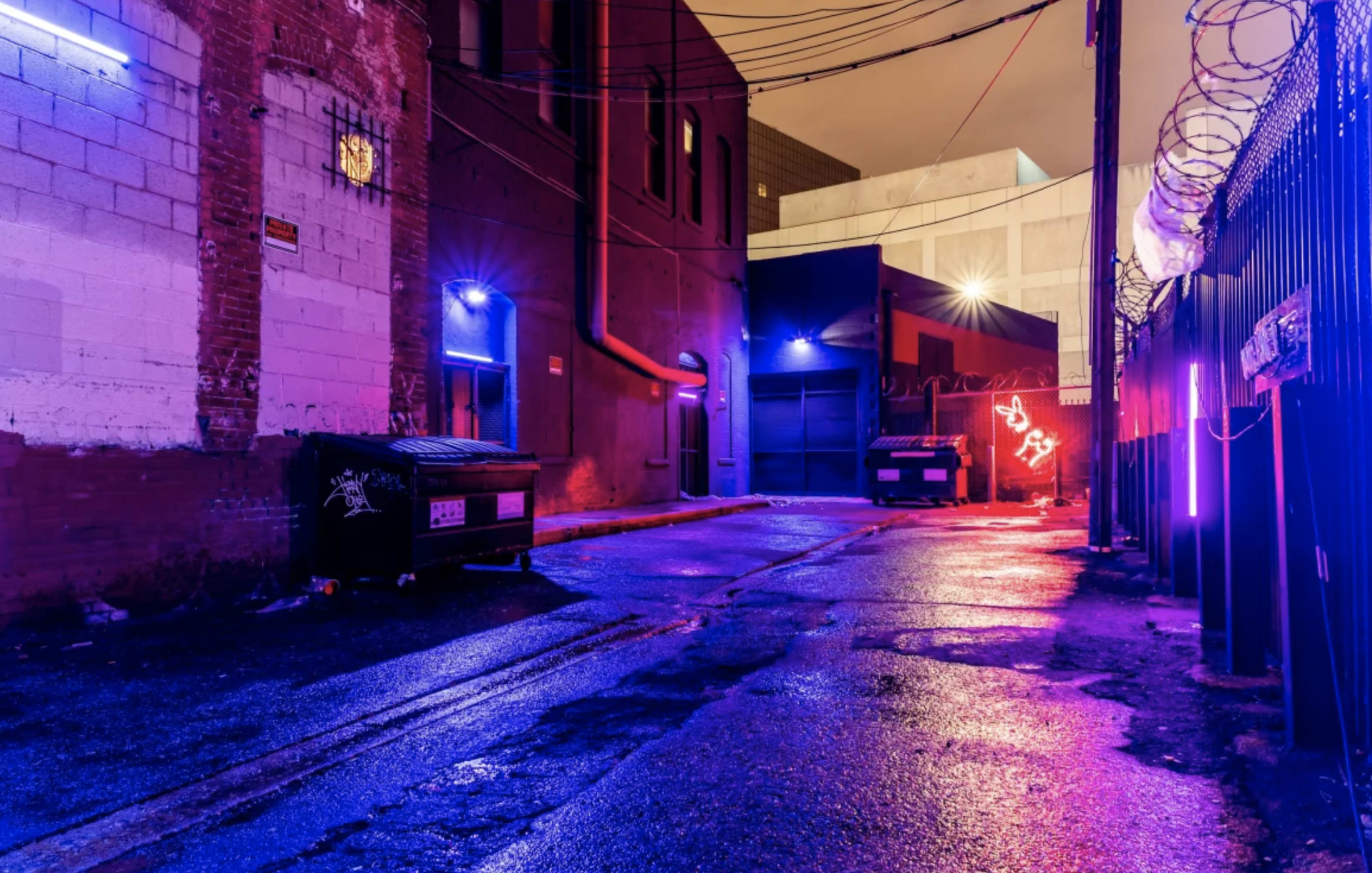 A wet alleyway at night, featuring a dumpster, a parked car, and illuminated signs casting colorful lights along the brick walls.