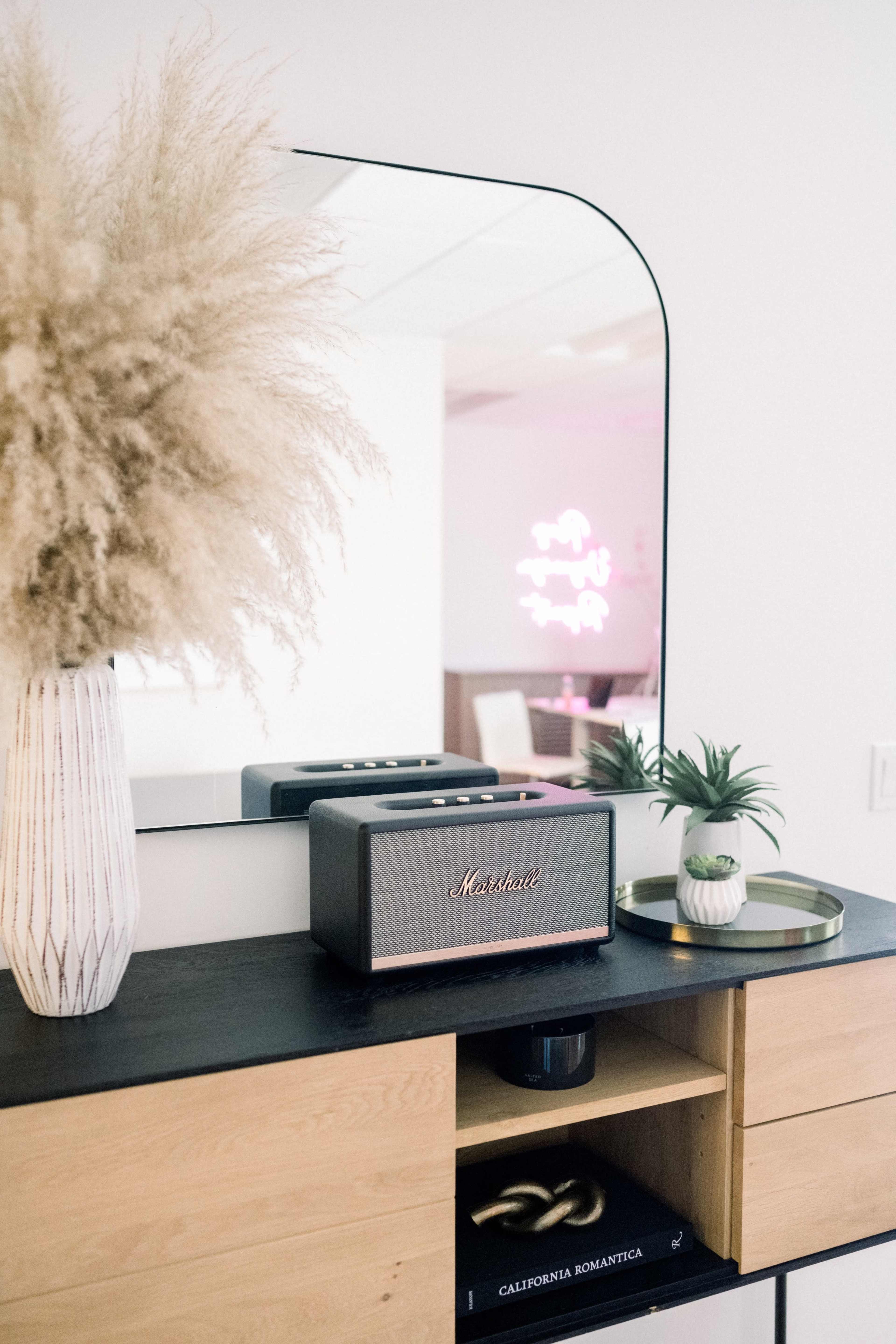 A stylish console table features a Marshall speaker, a decorative plant, a vase with pampas grass, and a round tray, all reflected in a large mirror.