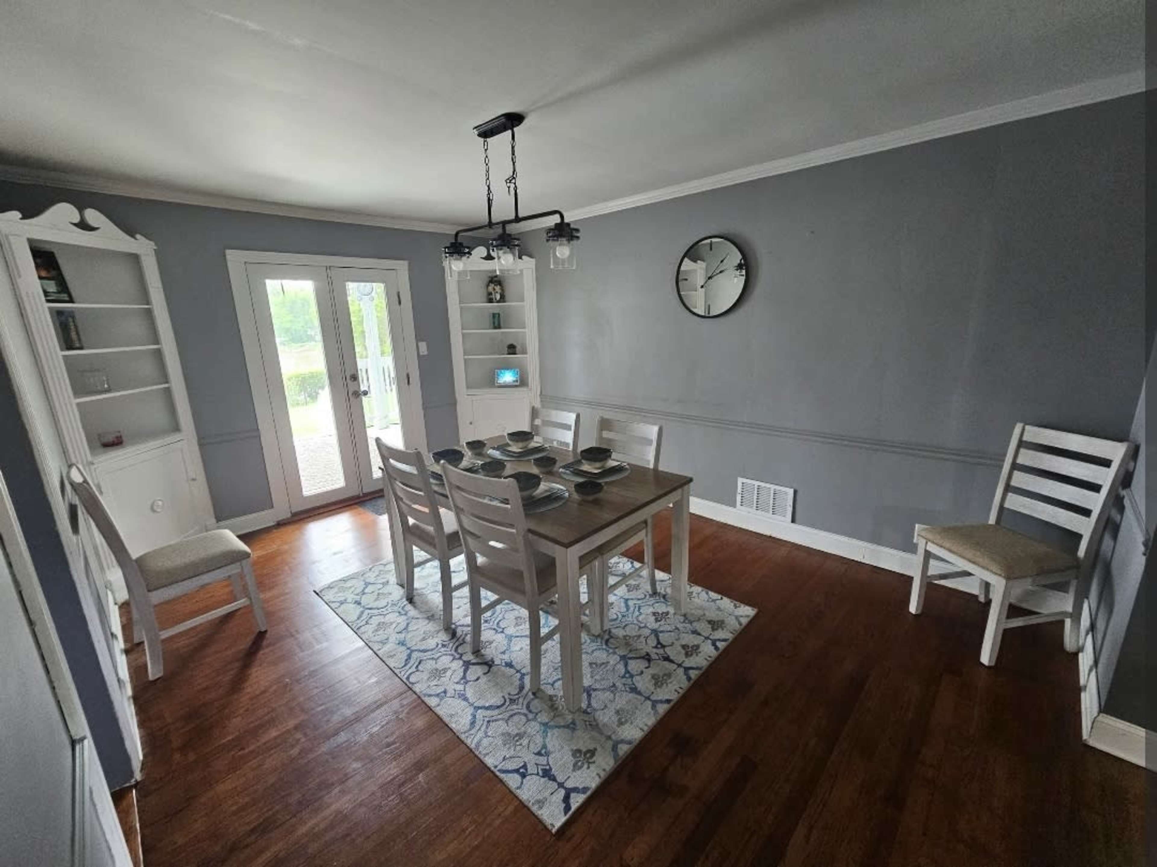 A dining room features a rectangular table set for six, surrounded by white chairs, with a clock and a cabinet against the gray walls.
