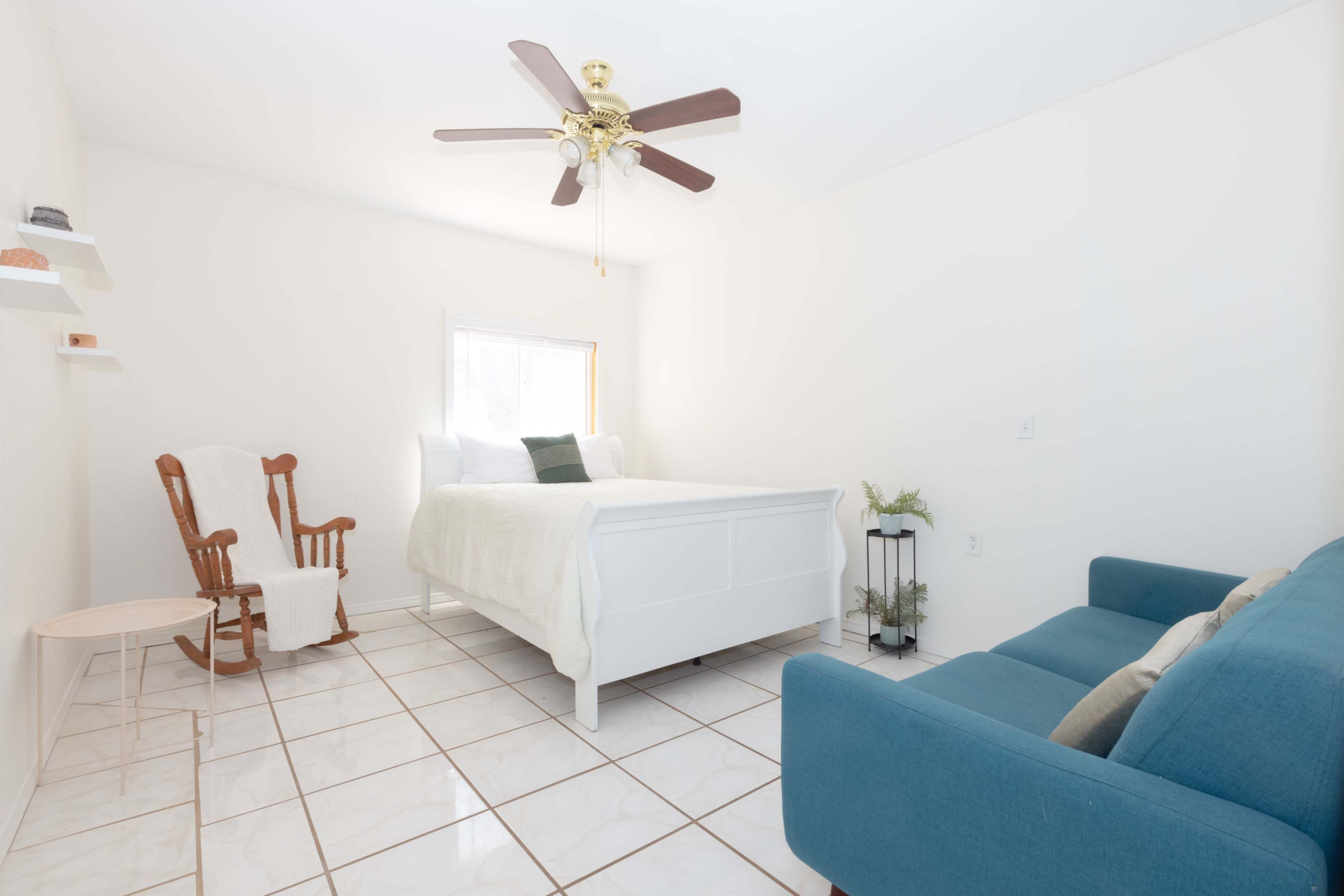 A bright, minimalist bedroom featuring a white bed, a rocking chair, and a blue sofa on tiled flooring.