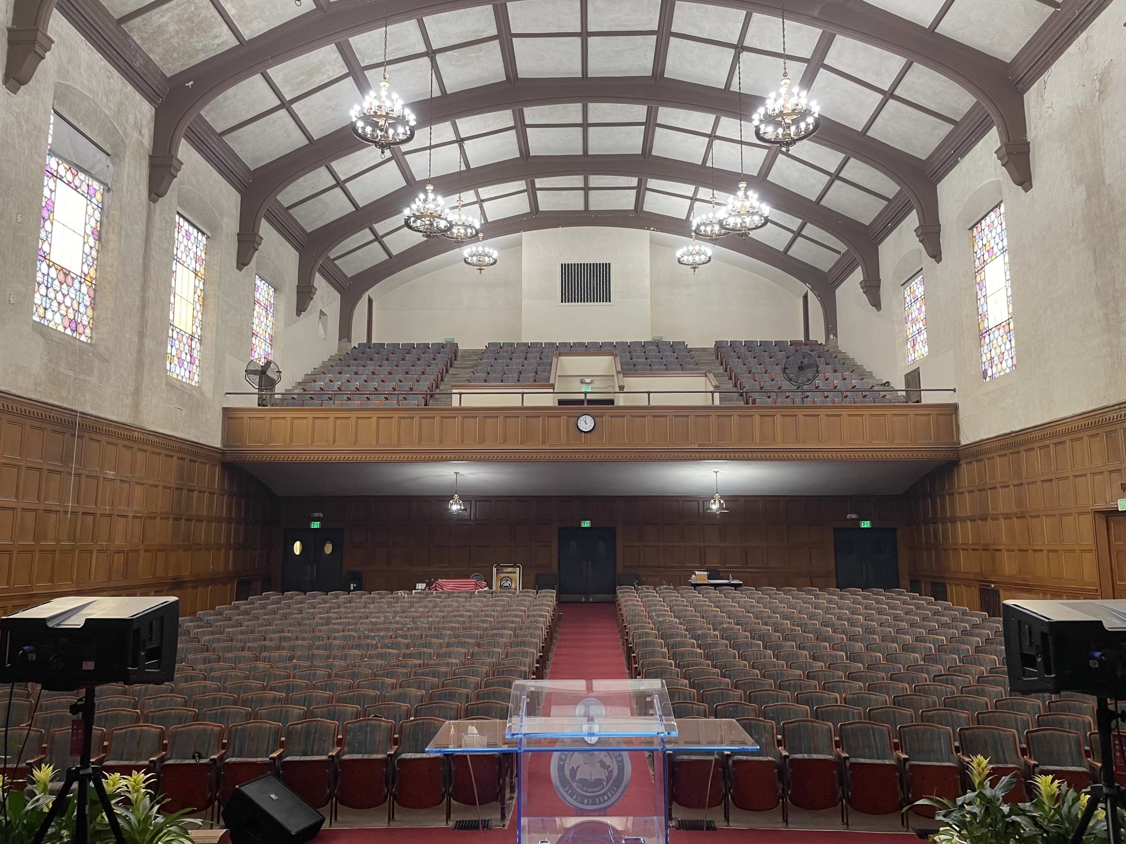The image shows a spacious, empty auditorium with rows of red-upholstered seats, a wooden stage in the foreground, and stained glass windows lining the walls.