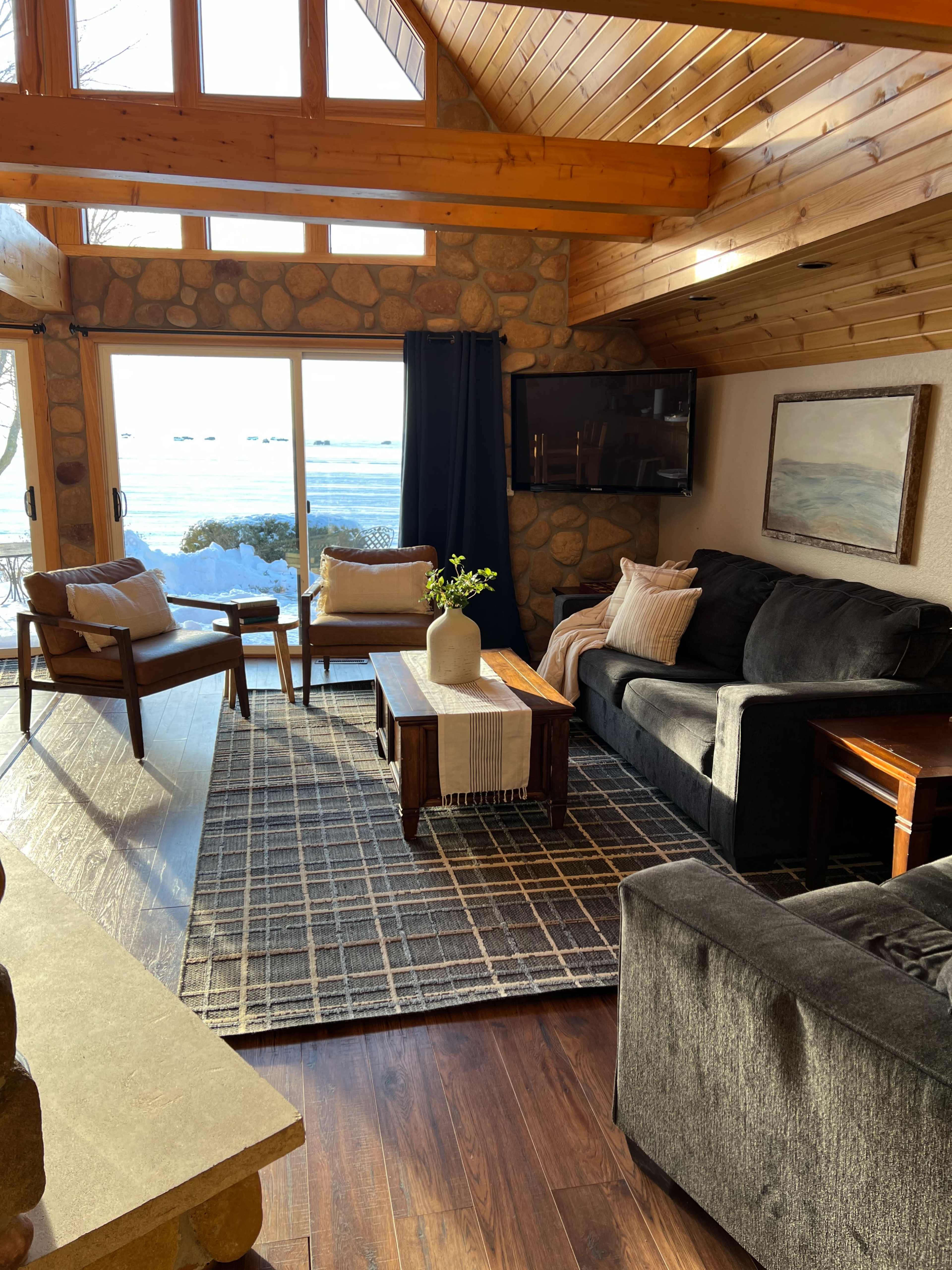 A cozy living room features a stone wall, wooden beams, a dark sectional sofa, and a coffee table with a vase of flowers, all overlooking a snowy landscape through large windows.