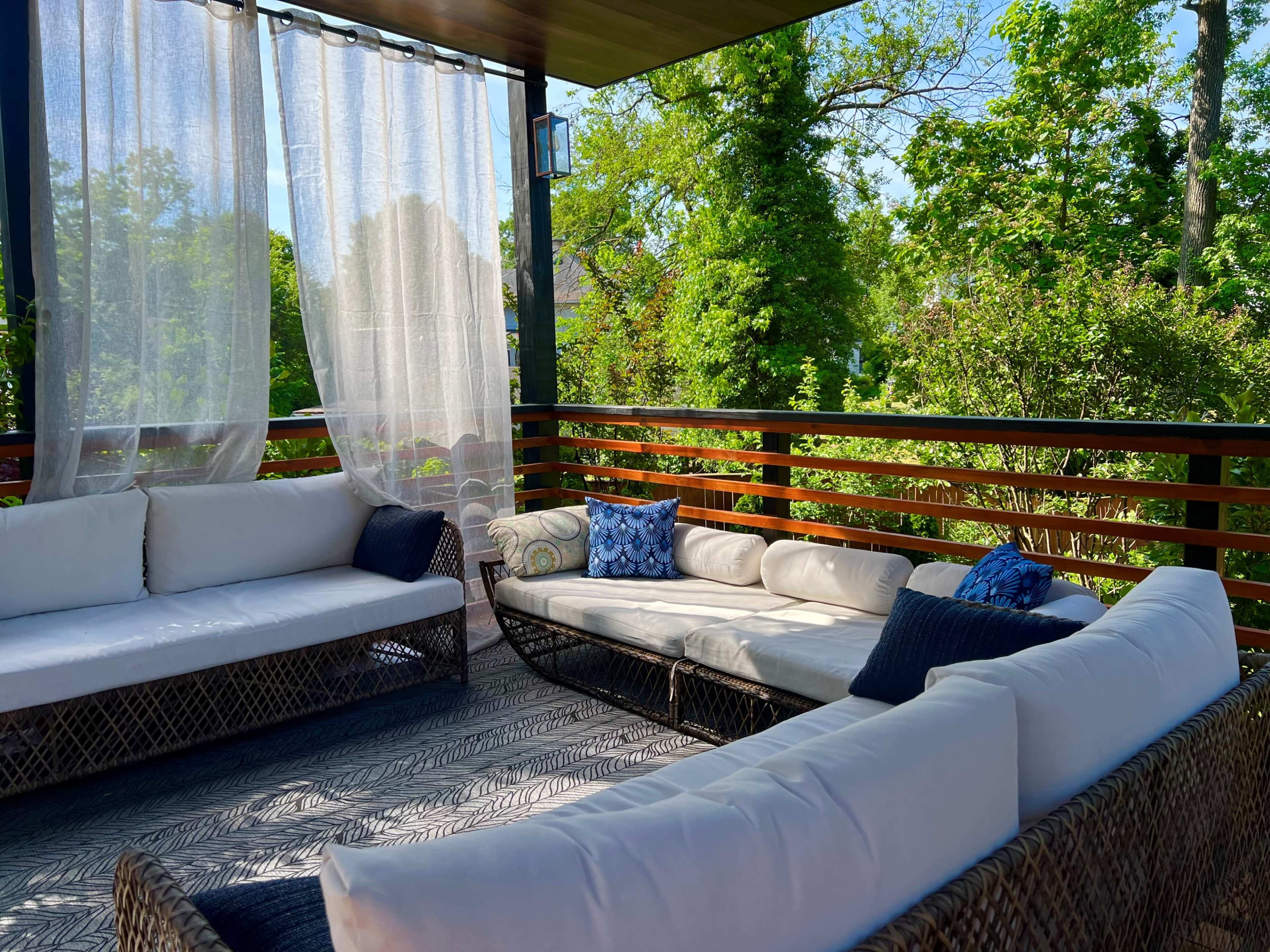 A cozy outdoor seating area features a wicker sofa set with white cushions, surrounded by lush greenery and partially draped sheer curtains.