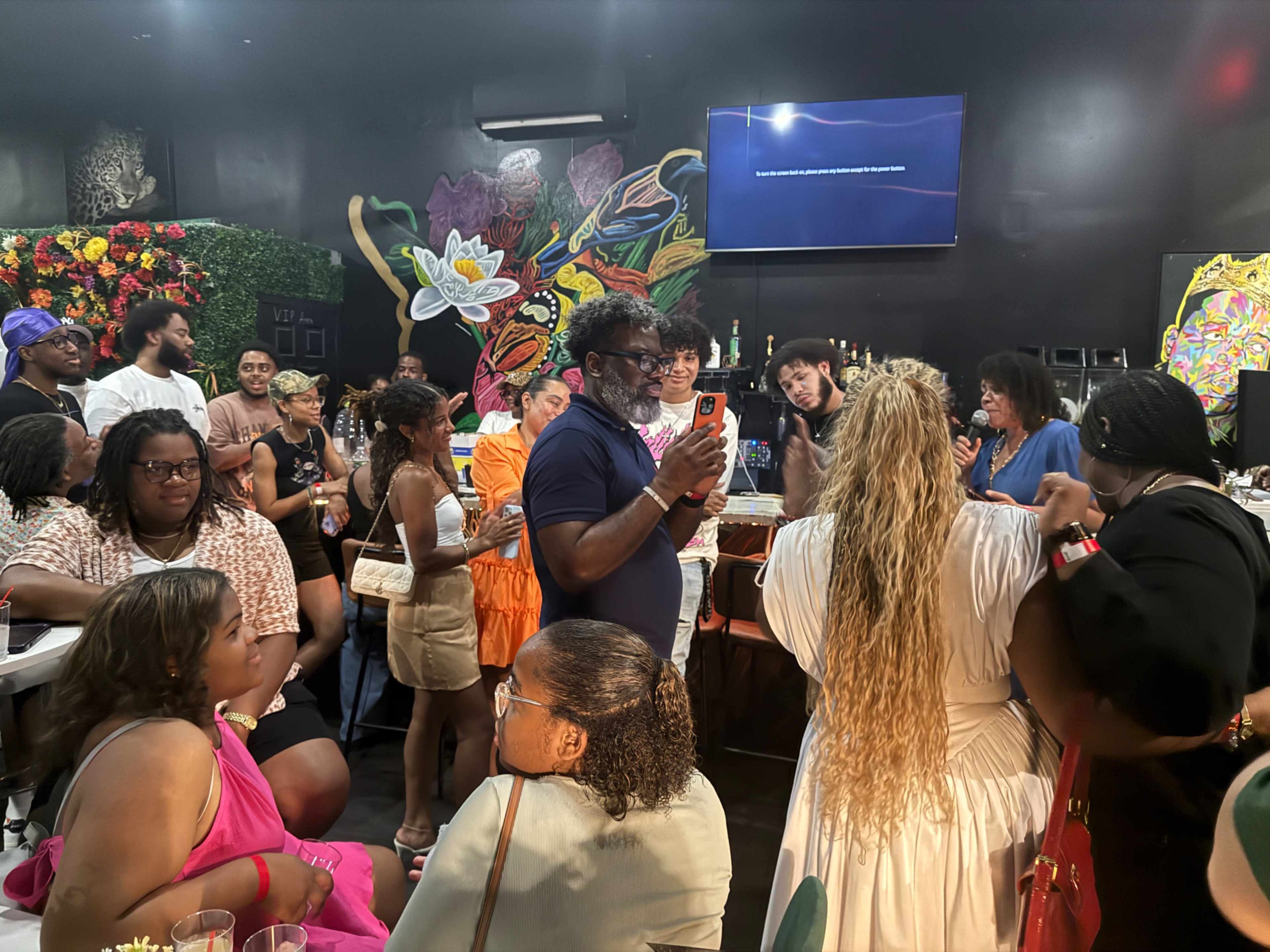 A crowd of people gathers in a lively bar setting, surrounded by colorful art and floral decorations.