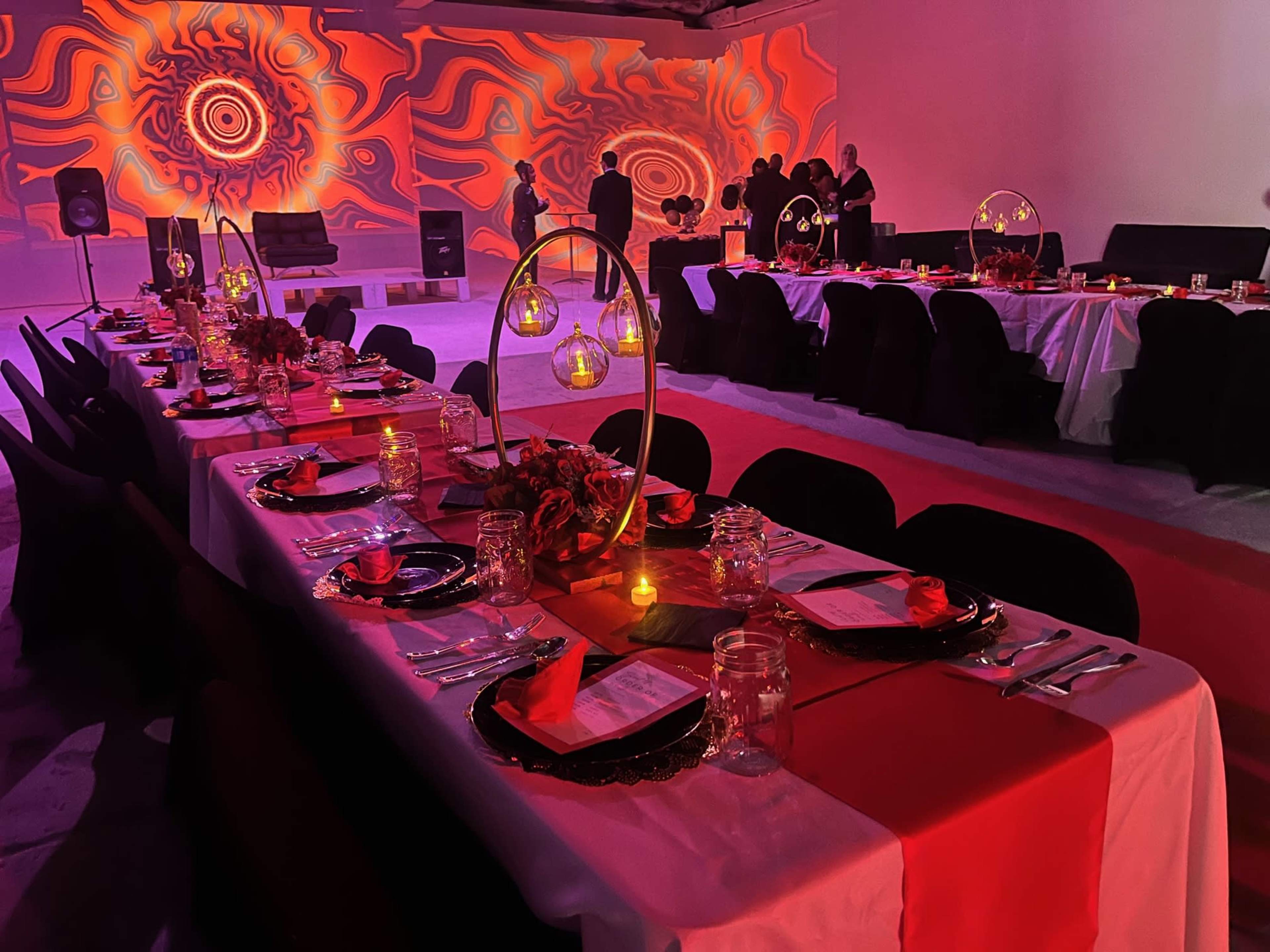 A long banquet table is elegantly set with plates, glasses, and decorative centerpieces, while a vivid red and orange light display illuminates the background and guests mingle in the distance.
