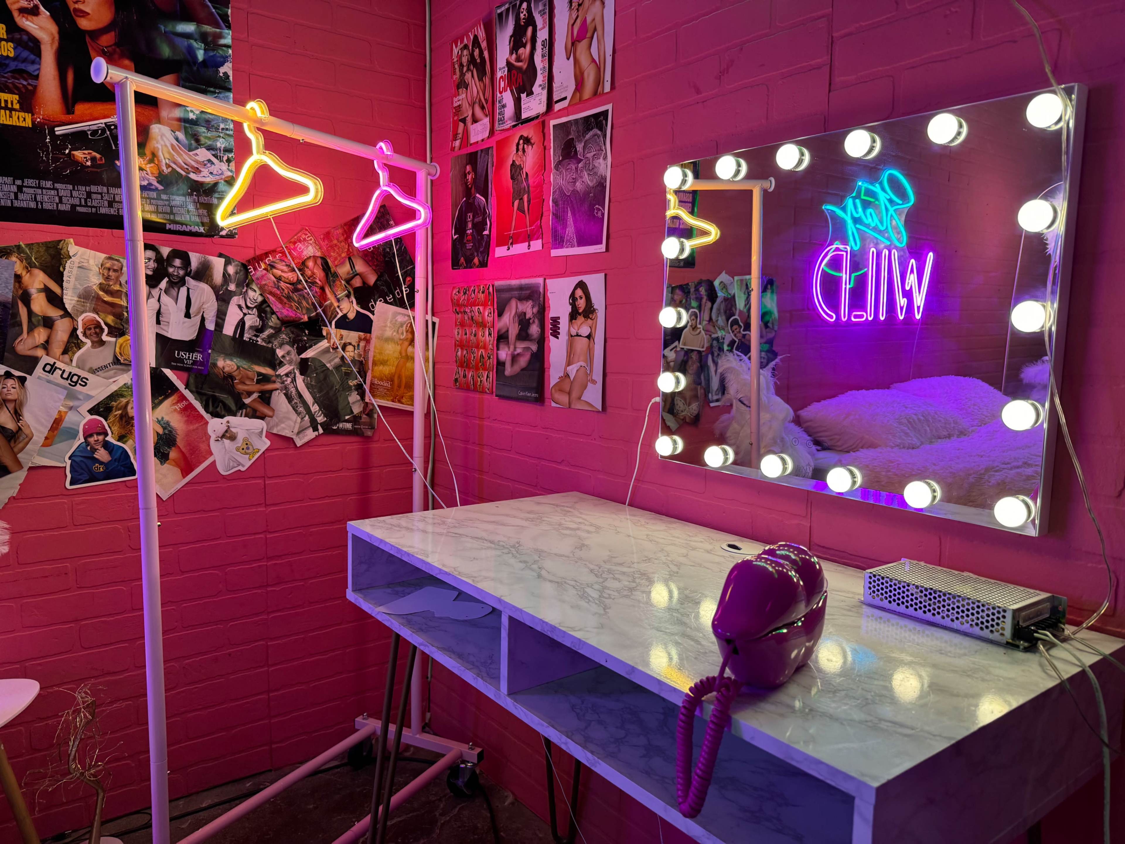 A pink-walled room with a marble desk, a mirror framed with lights, a neon sign, and a clothing rack with neon hangers.