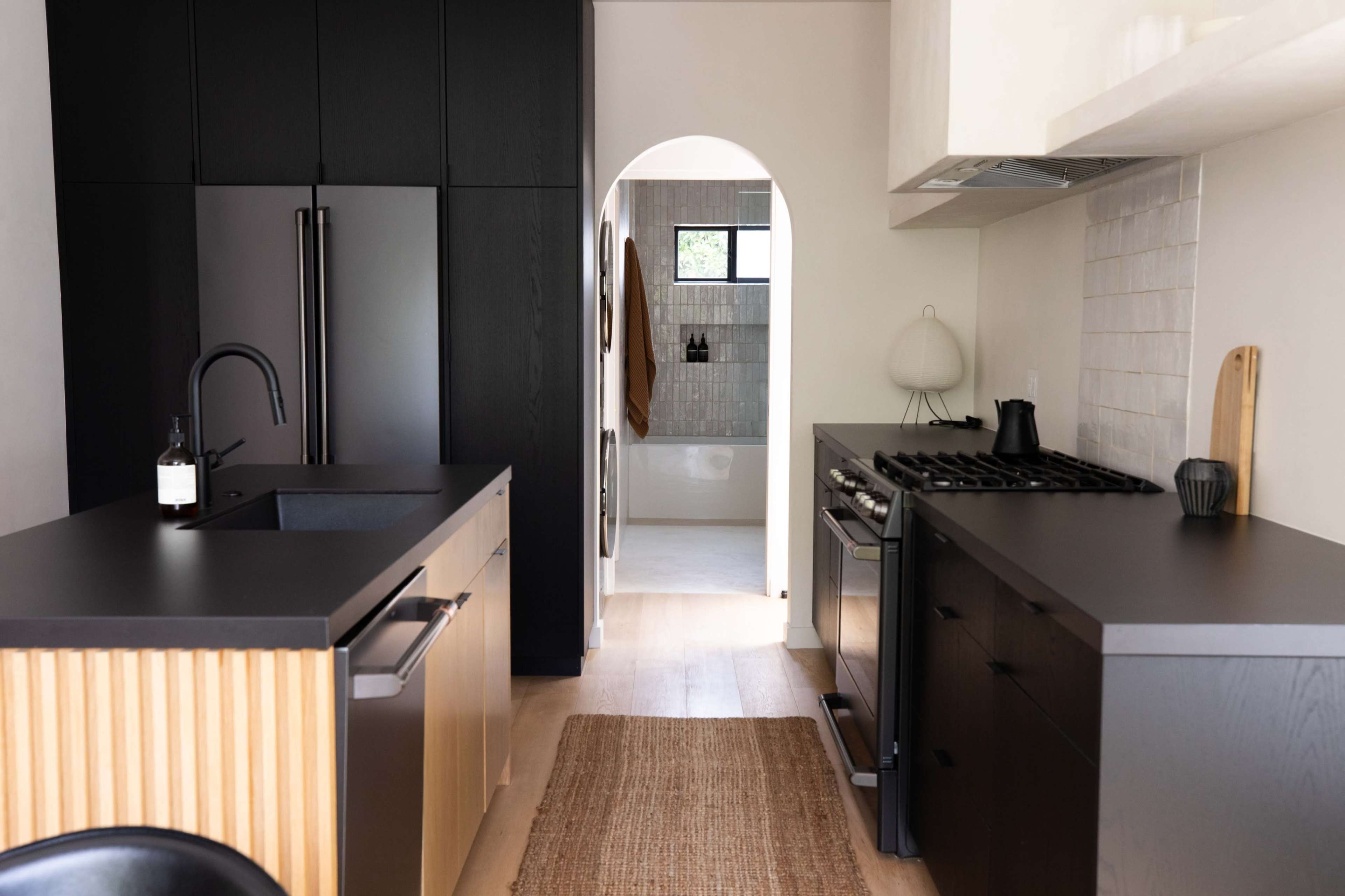 A modern kitchen features black cabinetry, a stainless steel stove, and a doorway leading to a bathroom.