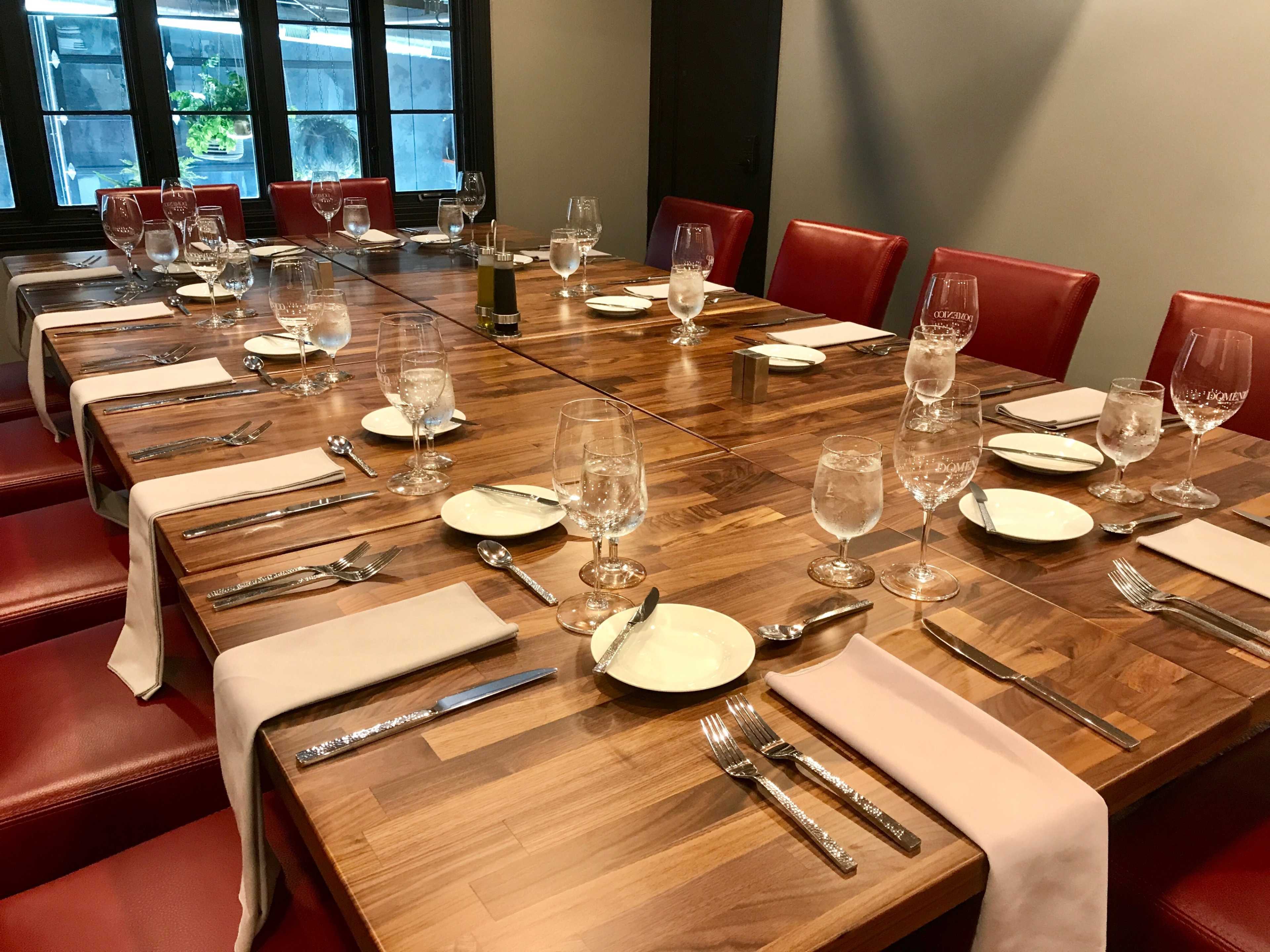 A long wooden dining table is set with plates, glasses, and utensils in a room with red chairs and large windows.