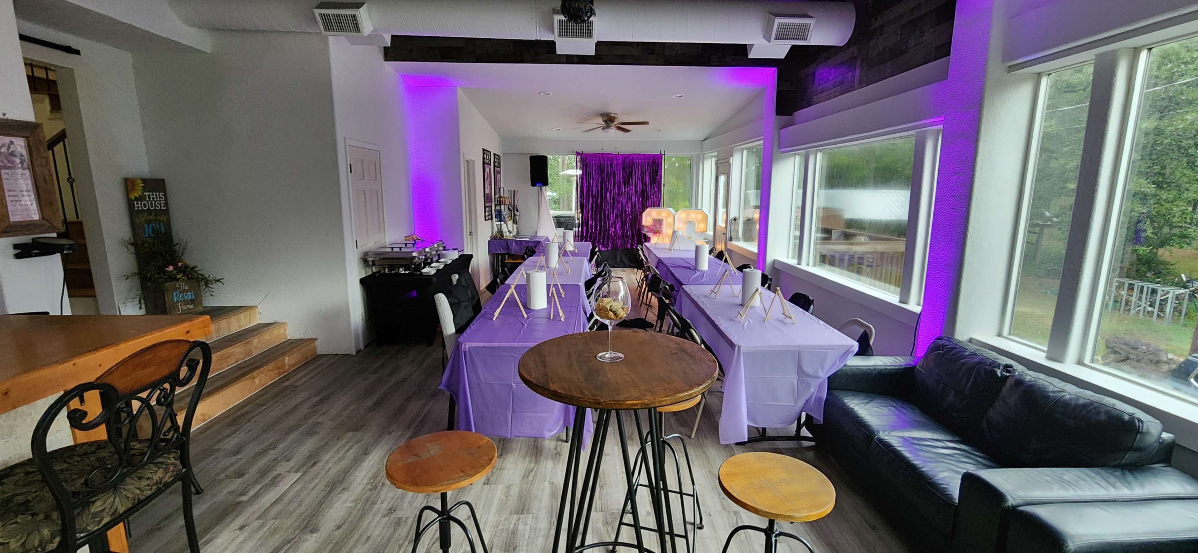 The image shows a modern event space set up for a gathering, featuring tables covered with purple linens, a casual seating area with a black sofa, and ambient purple lighting.