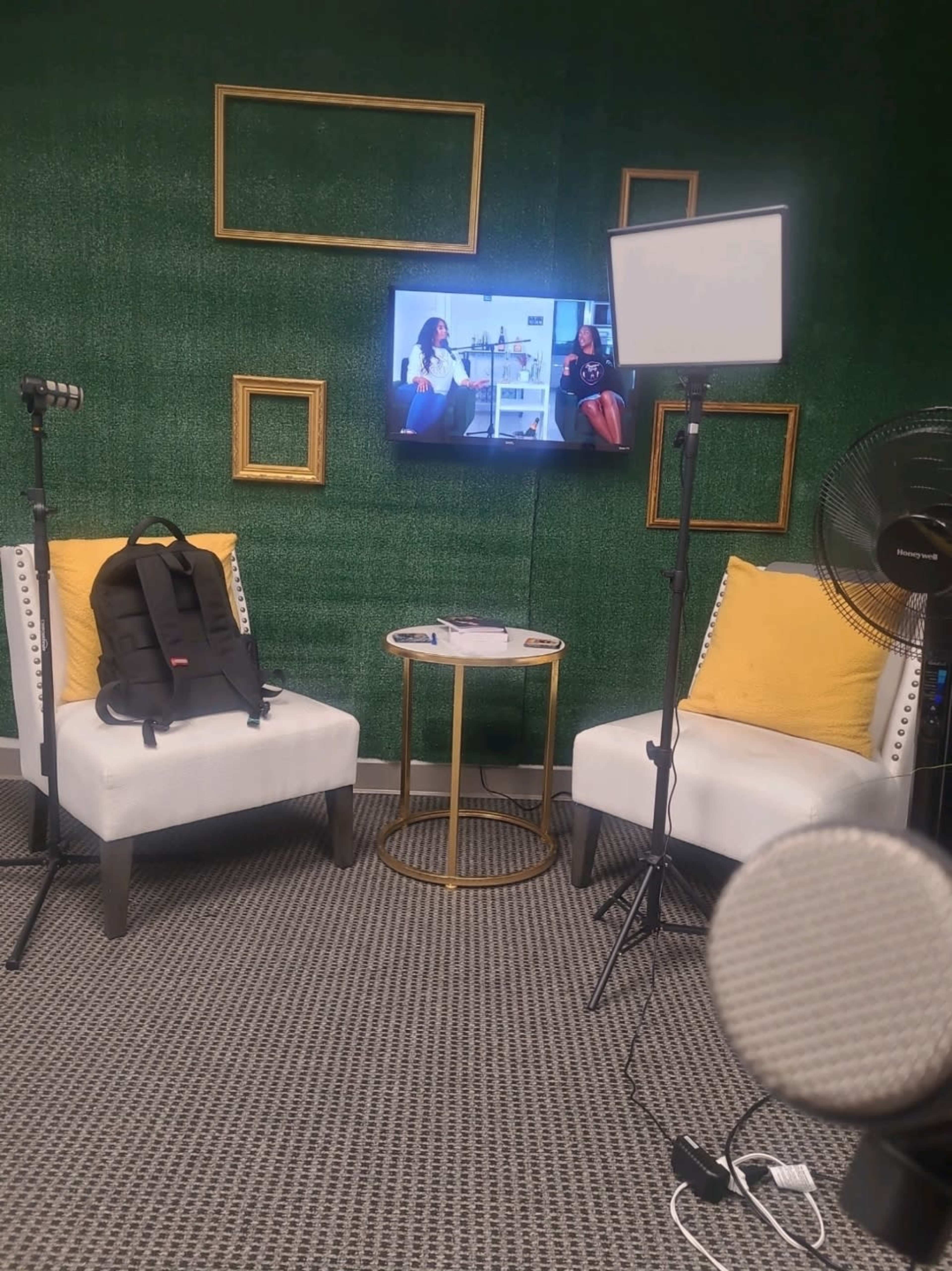 A green-walled studio setup features two white chairs with yellow cushions, a small side table, a television displaying two people, and various recording equipment.