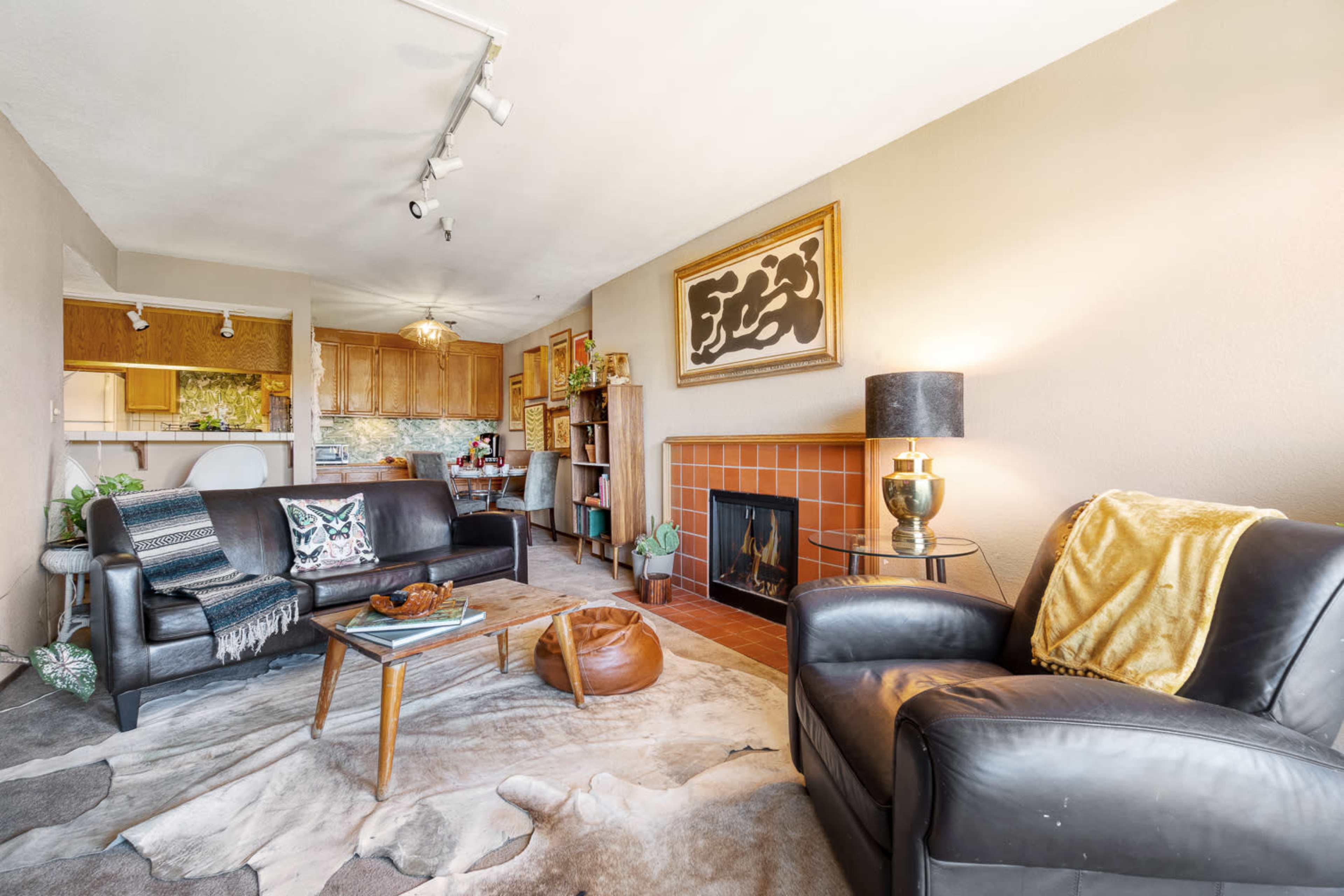 A cozy living room featuring a fireplace, a leather armchair, a coffee table, and a kitchen in the background.