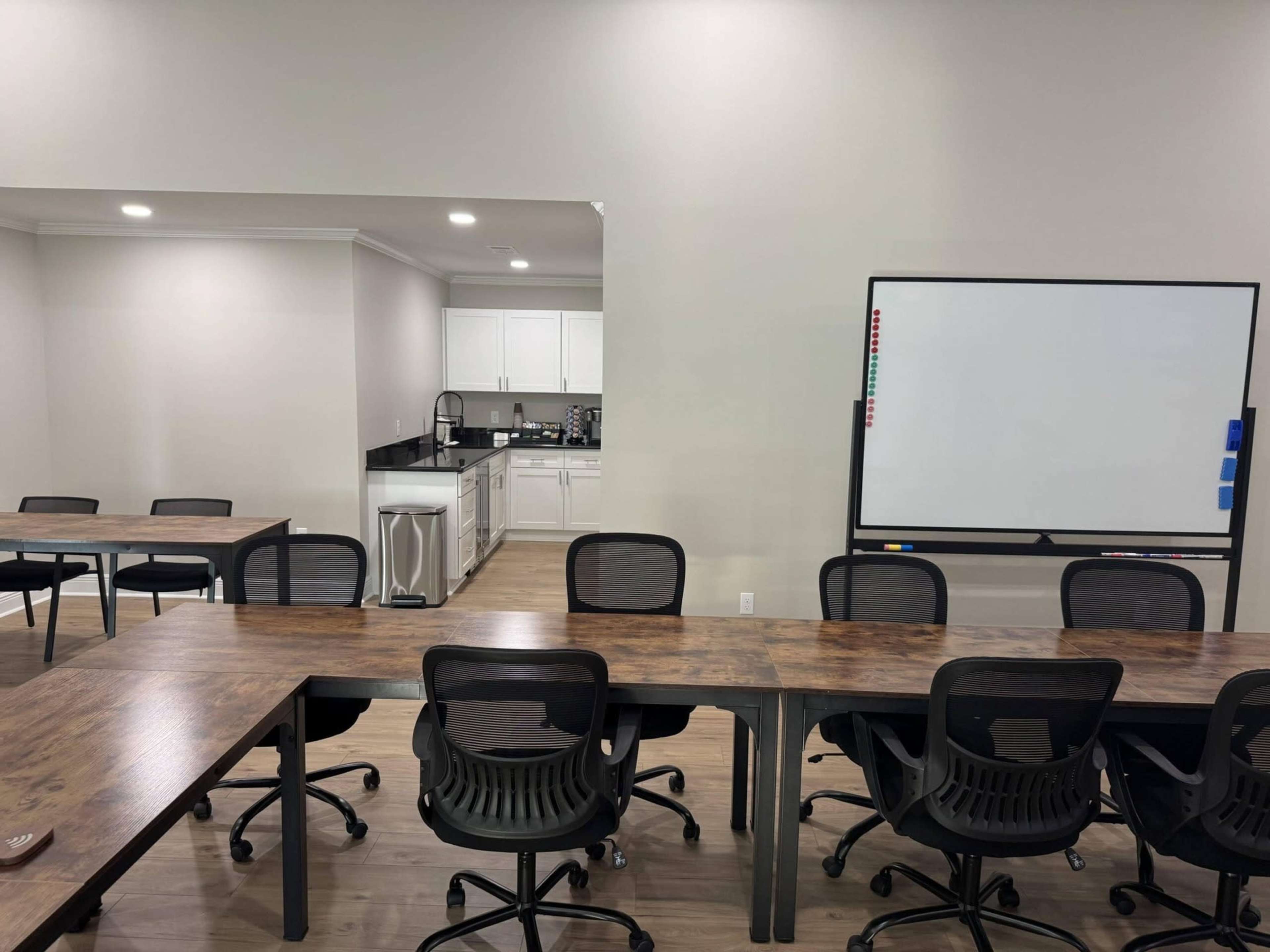 The image shows a conference room with several long wooden tables and black office chairs, adjacent to a kitchen area featuring white cabinets and a whiteboard.