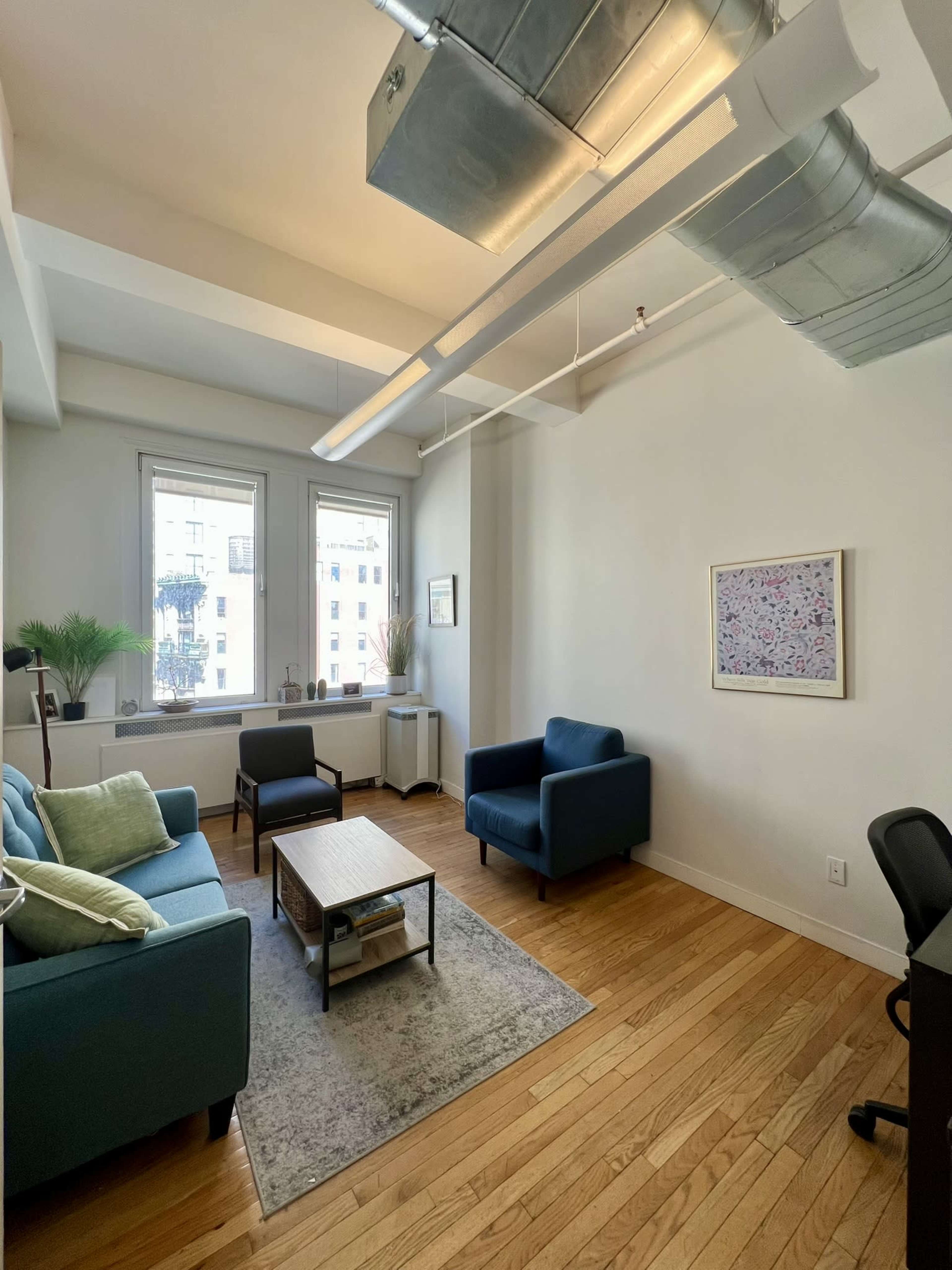 The image shows a bright office space with two blue couches, a coffee table, and a desk, featuring large windows that let in natural light.