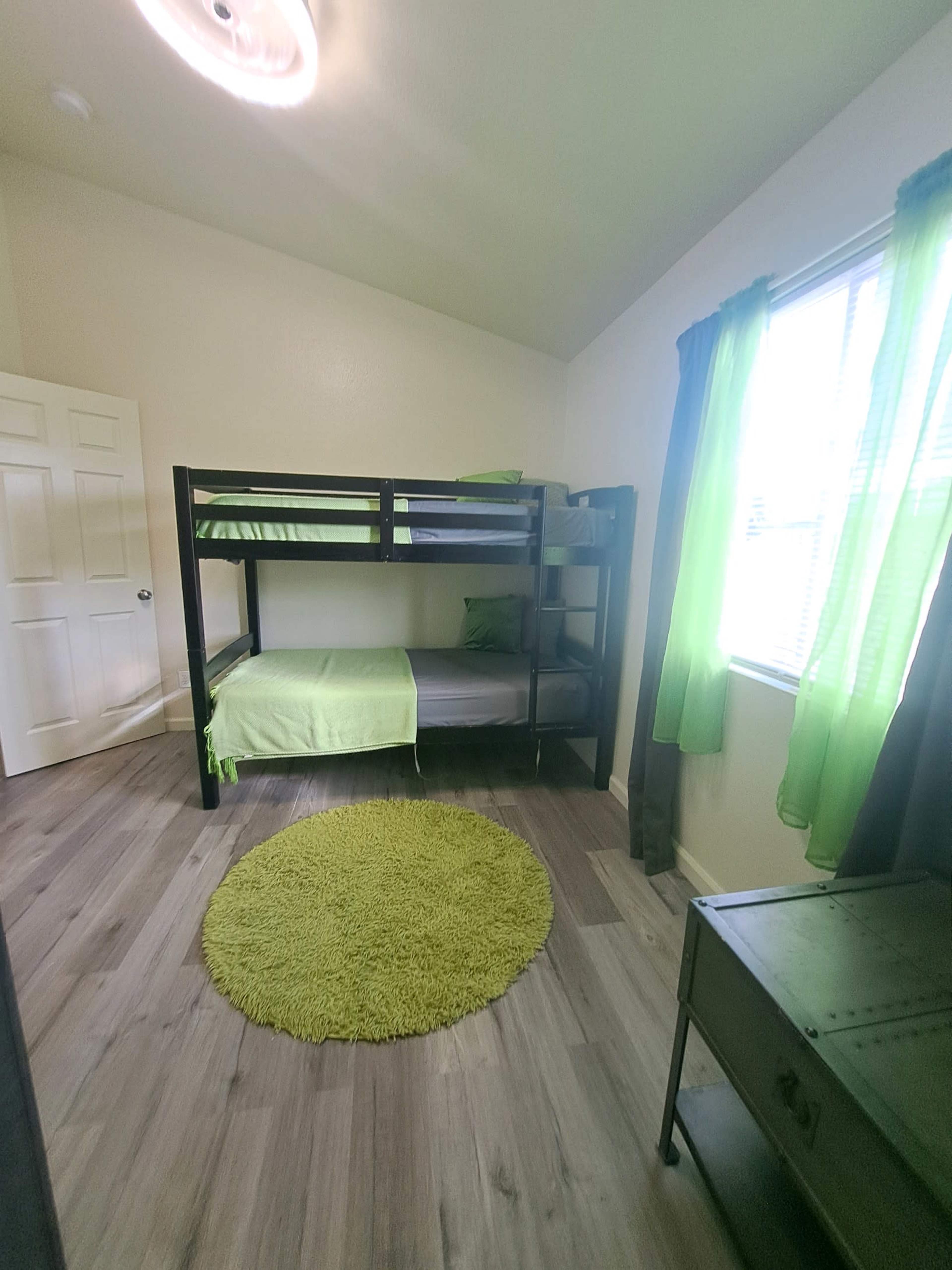 The image shows a small bedroom featuring a black bunk bed with green bedding, a round green rug on the floor, and large windows covered with green curtains.