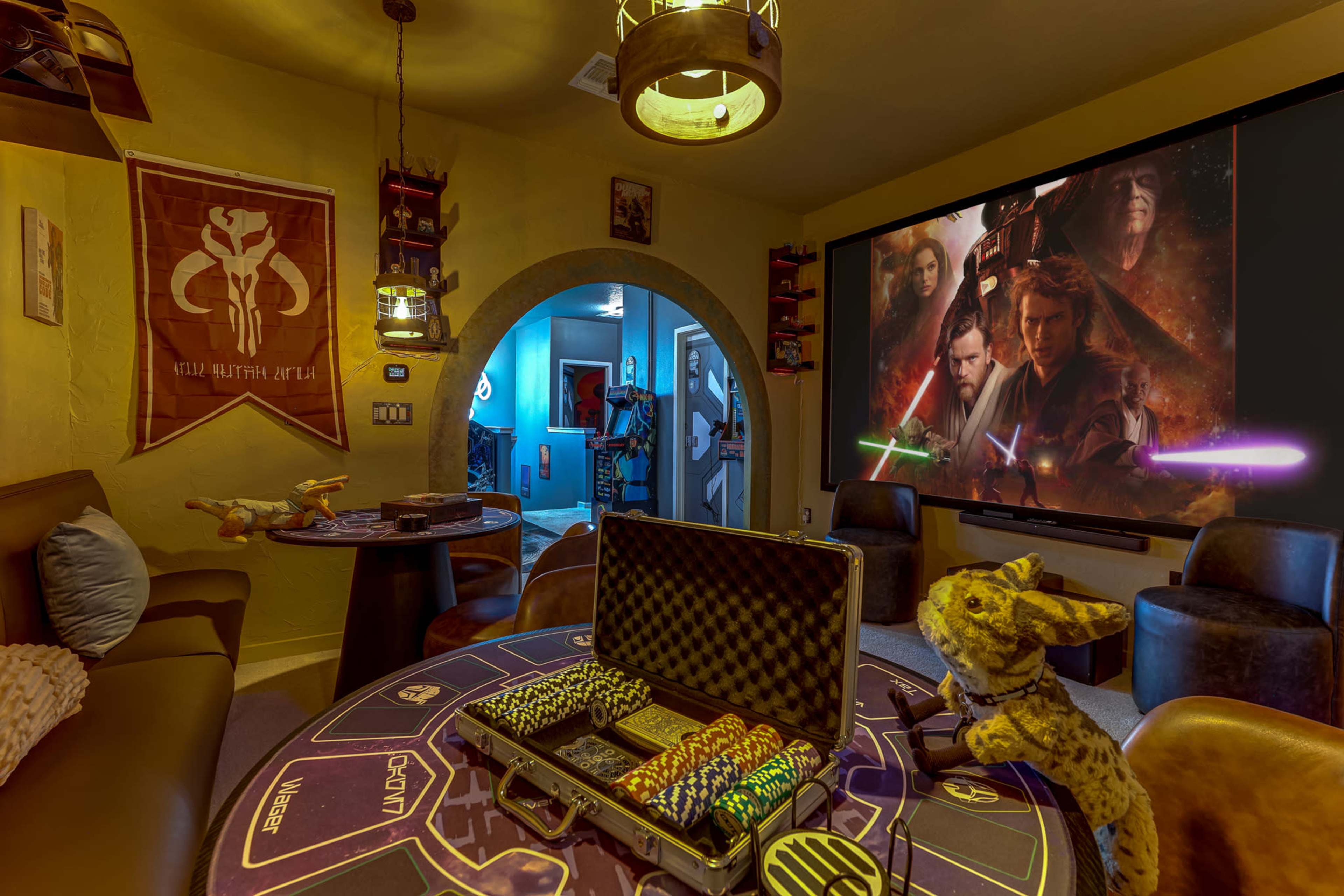 The image shows a themed gaming room featuring a large wall screen displaying characters with lightsabers, along with a gaming table, chairs, and decorative elements related to the Star Wars franchise.