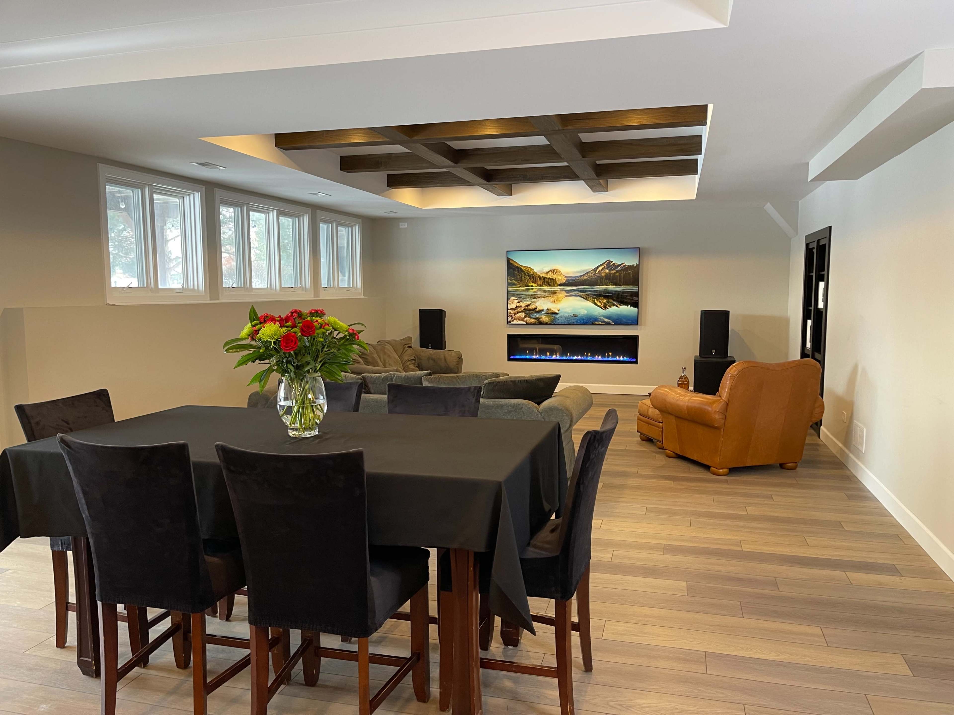 A modern living space features a dining table with black chairs, a sectional sofa, a brown leather armchair, and a wall-mounted television above a scenic painting, all arranged under a coffered ceiling.
