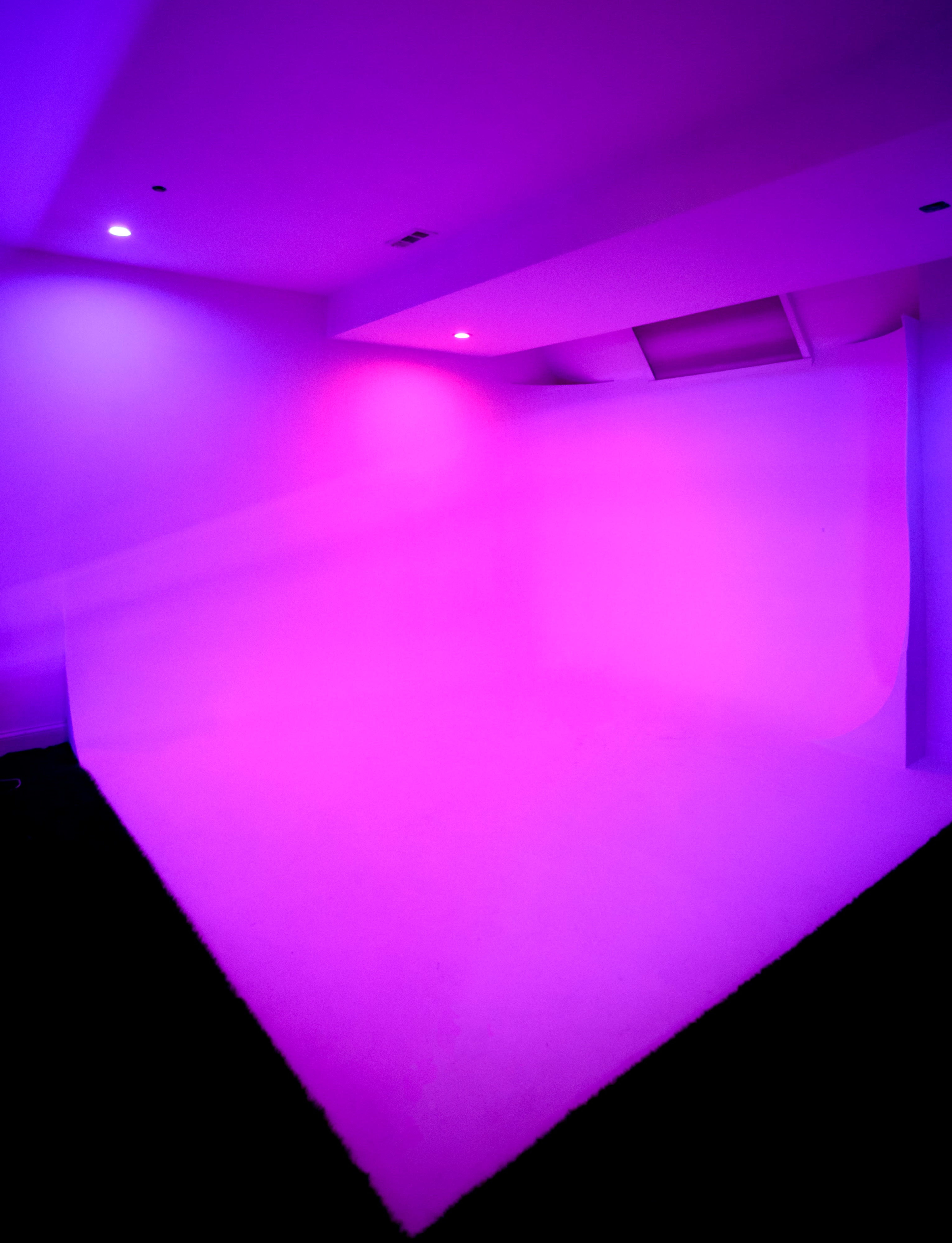 A minimalist studio space is illuminated with purple lighting, featuring a smooth white backdrop and a dark floor.