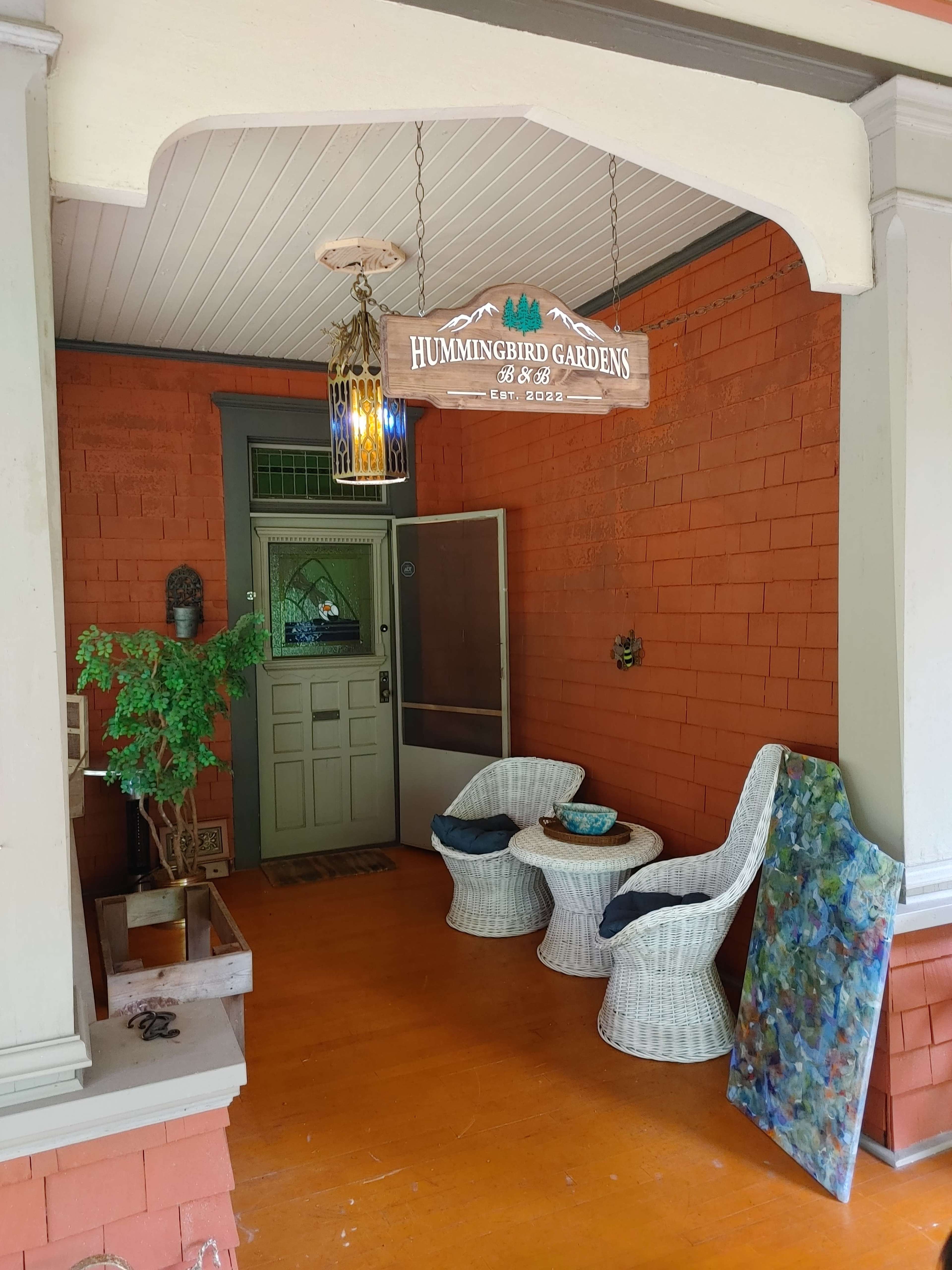 A front porch with a decorative sign reading "Hummingbird Gardens," two wicker chairs, a small table, and a colorful piece of art against the wall.